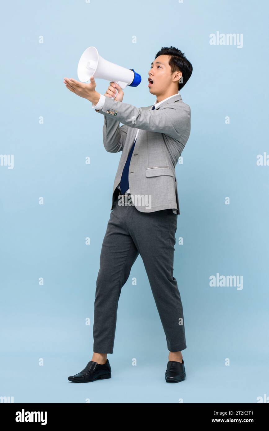 Young handsome Asian man in formal business suit holding megaphone and yelling in isolated light blue color studio background Stock Photo