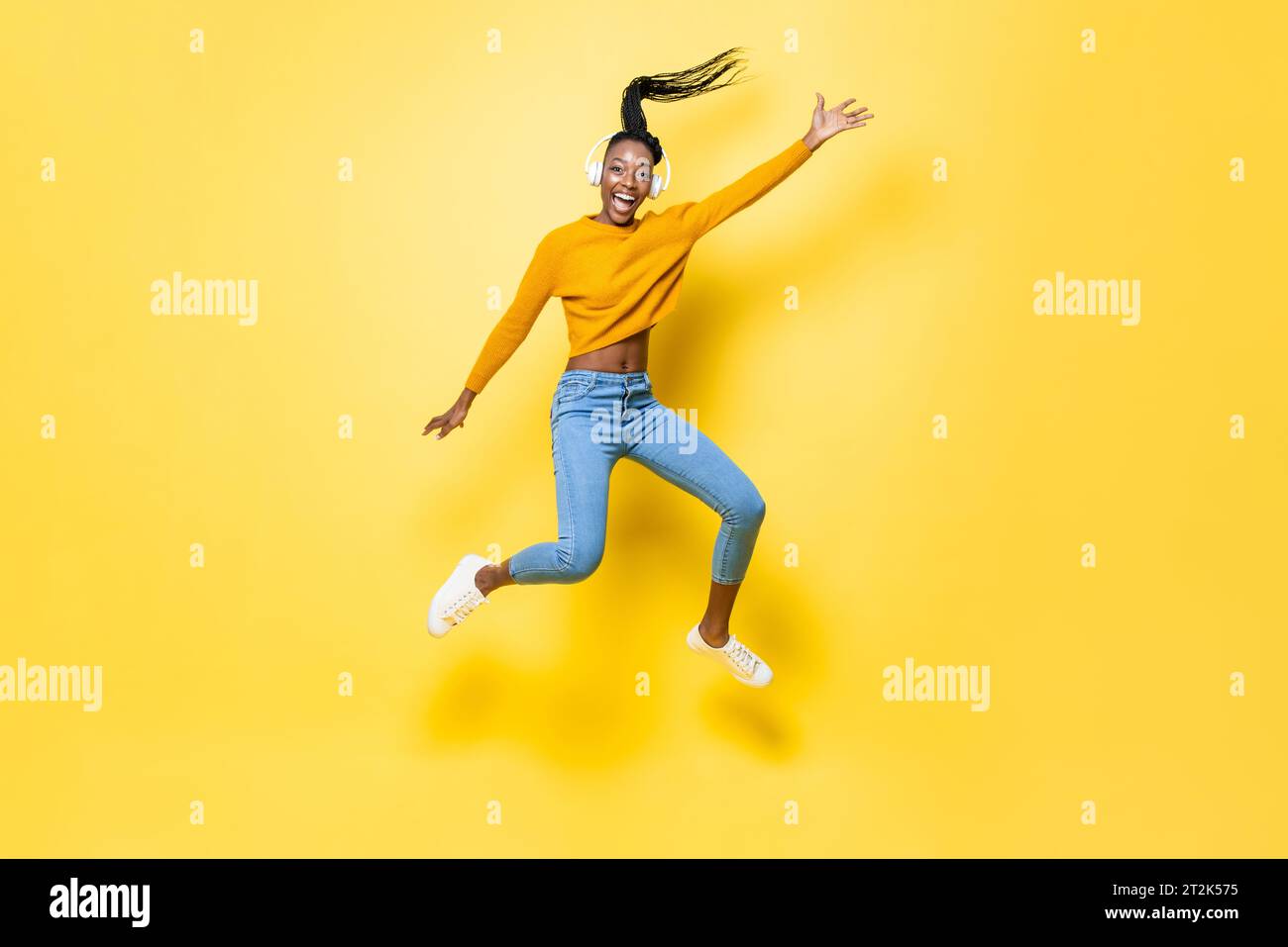 Positive enthusiastic young African American woman wearing headphones listening to music and jumping with hand up in yellow isolated studio background Stock Photo