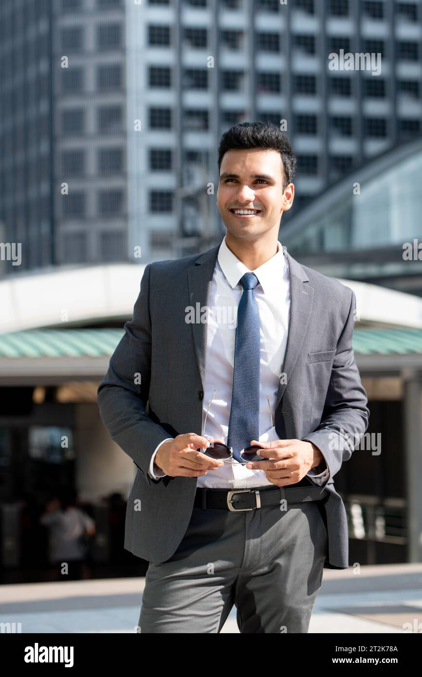Portrait of smiling elegant young Indian businessman wearing gray suit and holding sunglasses while standing against buildings in city Stock Photo