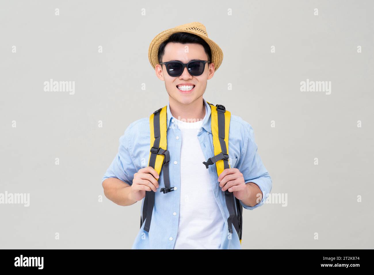 Porttrait of young smiling Asian tourist man with backpack wearing hat and sunglasses read for summer vacations, studio shot isolated in gray backgrou Stock Photo