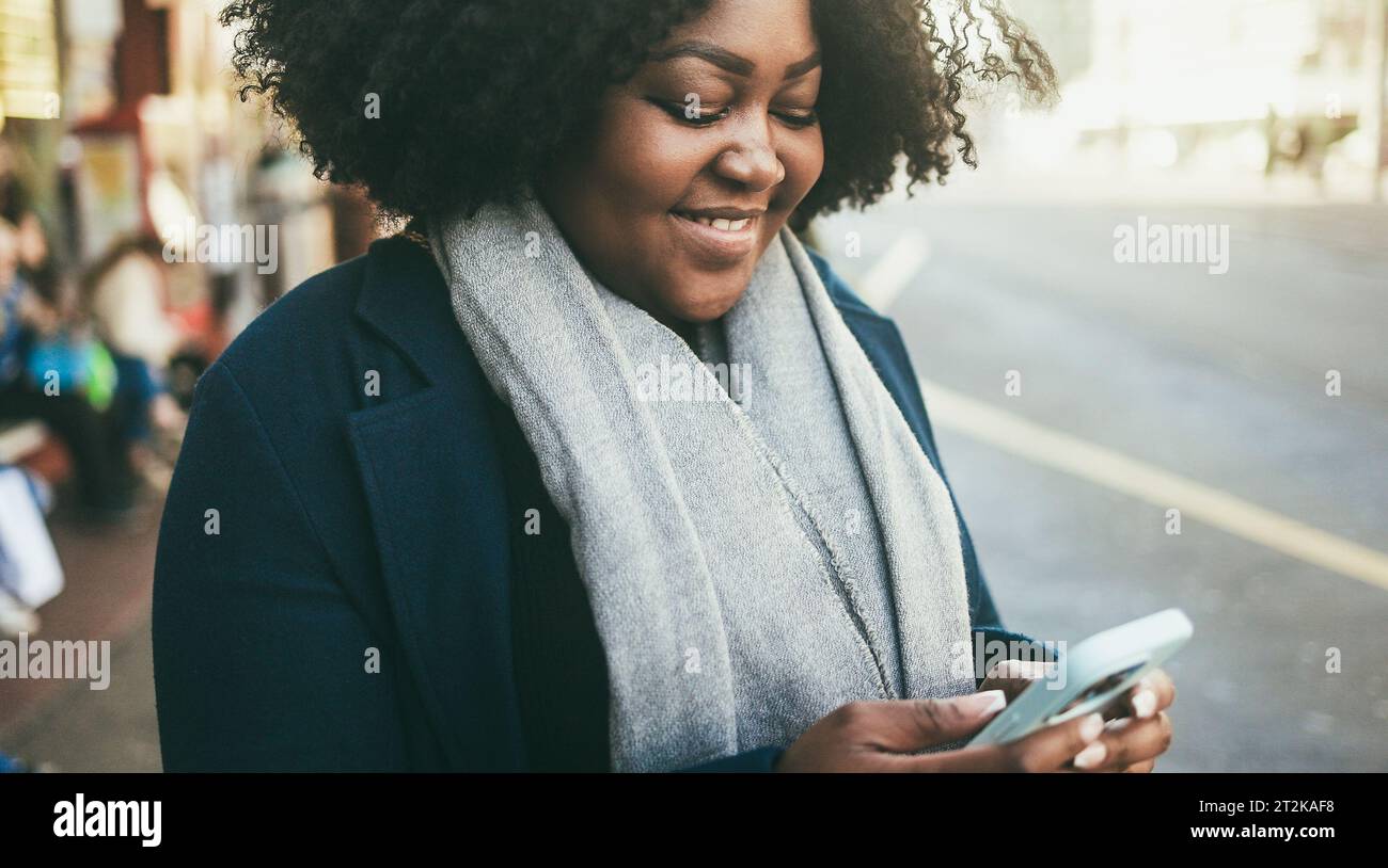 Curvy african woman using smartphone outdoor at bus station - City lifestyle and winter concept - Soft focus on face Stock Photo