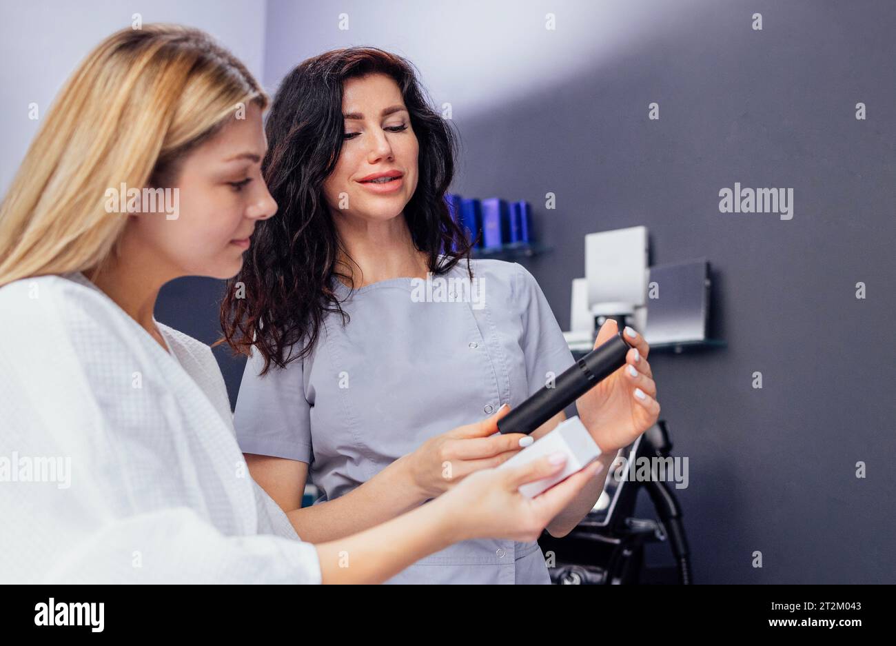 Pretty caucasian beautician communicates with her female client. Beauty master holds cosmetics in her hands and shows them to blod woman. Apparatus fo Stock Photo