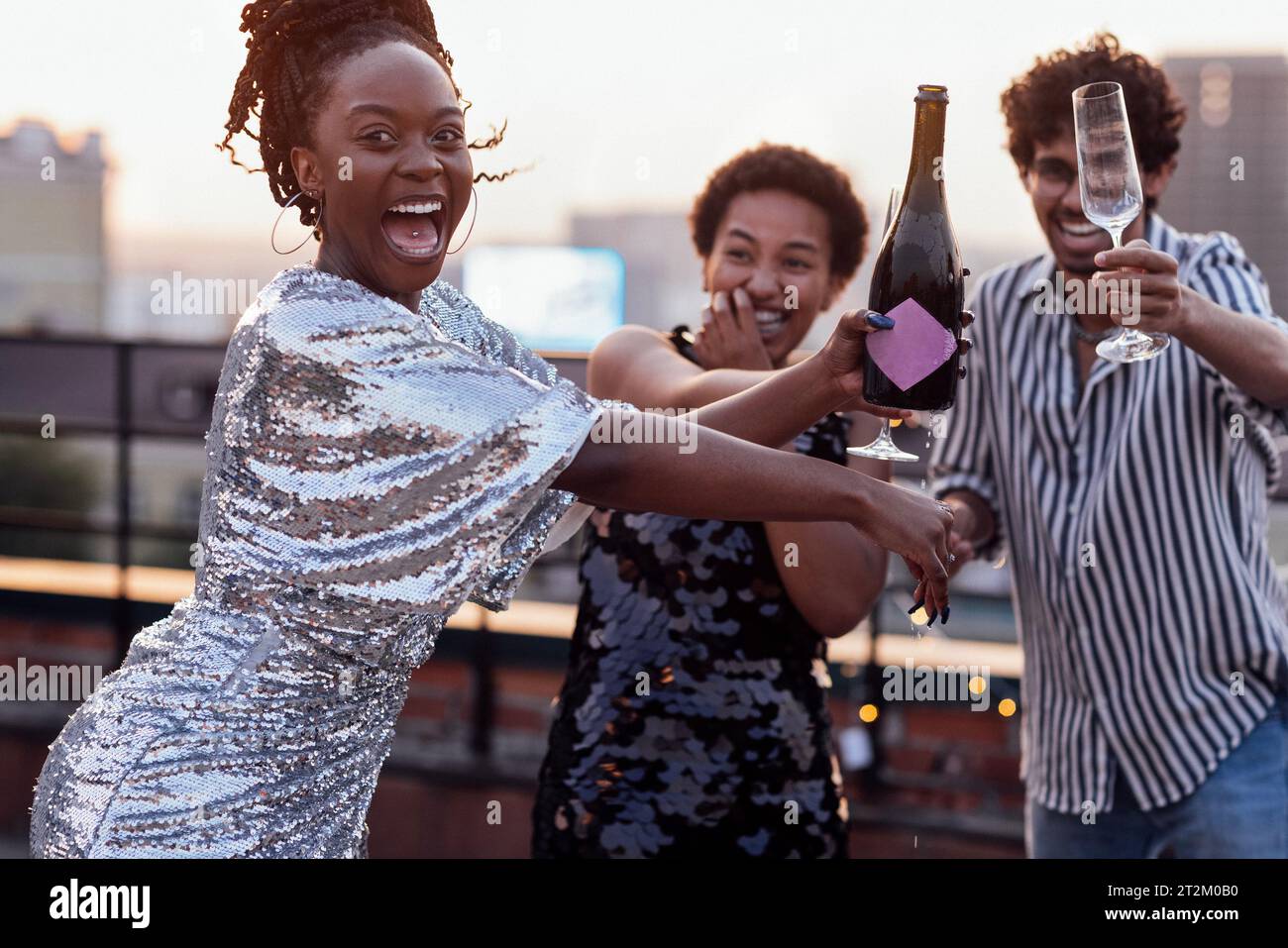 Dark-skinned girl pours champagne into glasses of her multinational friends. Cute happy people having fun at rooftop party. African women in elegant d Stock Photo