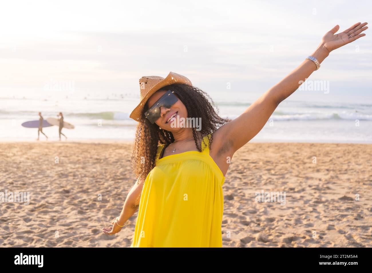 Mixed-raced happy woman wearing sunglasses and sun hat raising arms gesturing well-being on the beach Stock Photo