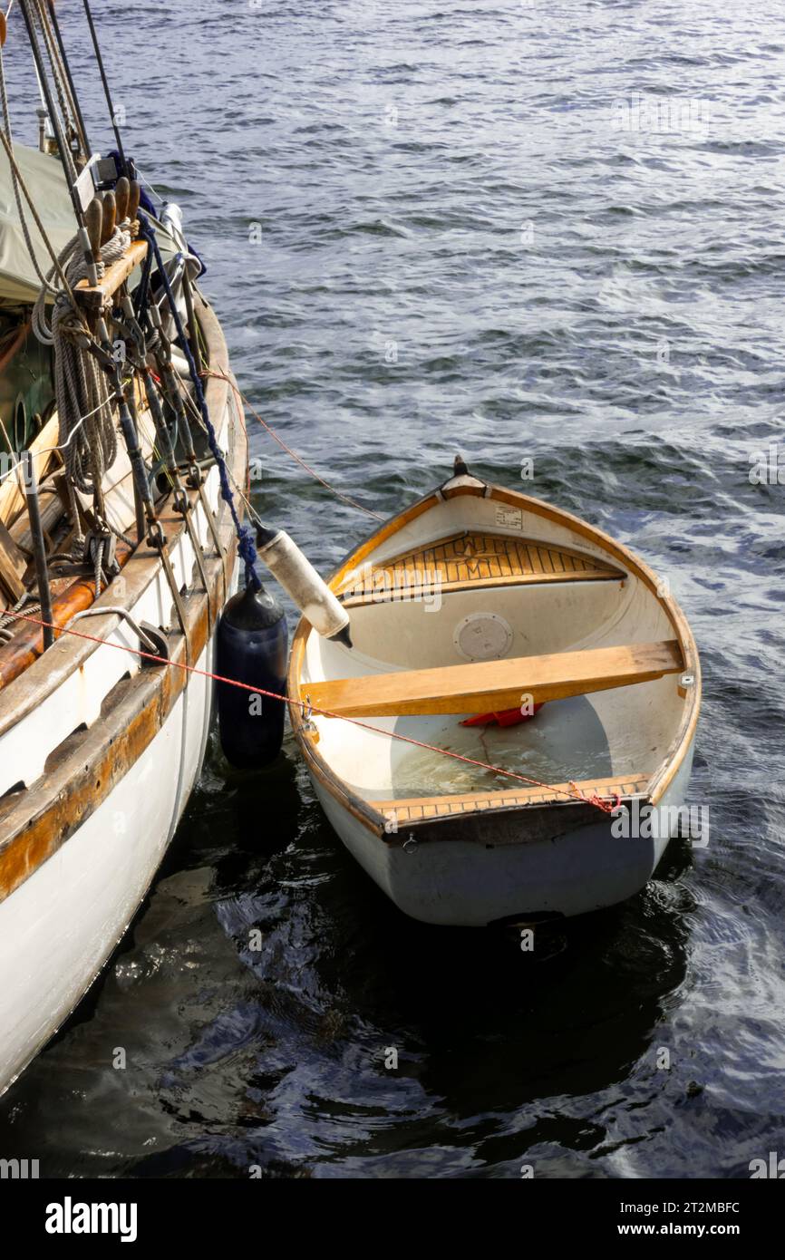 Small old wooden rowing boat moored with rope to a historical white sailboat as a rescue boat for nautical concept Stock Photo