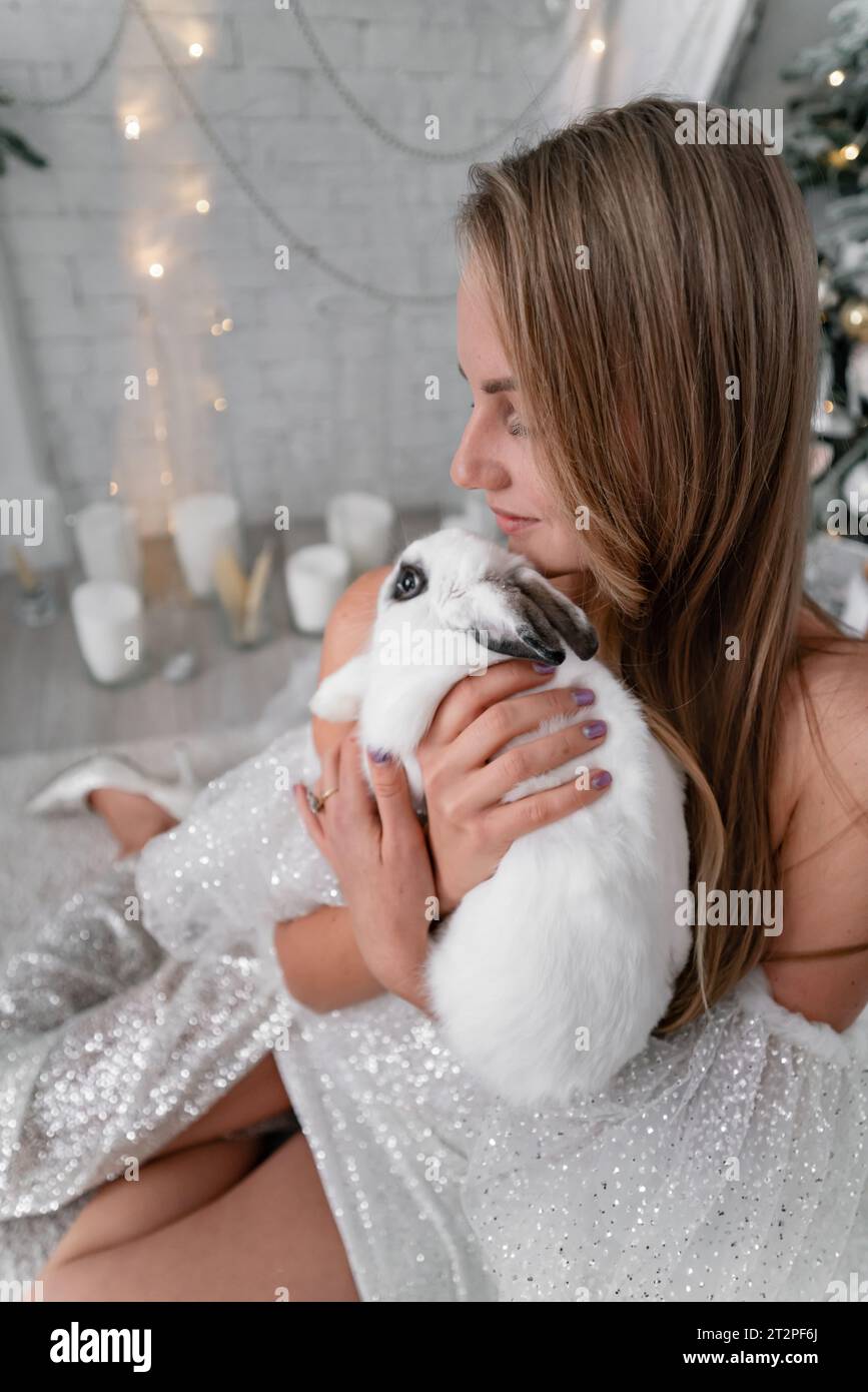 Woman holding a white rabbit symbol of the year 2023. Close-up of a beautiful young blonde woman holding a rabbit in a sparkly dress. She sits in a Stock Photo