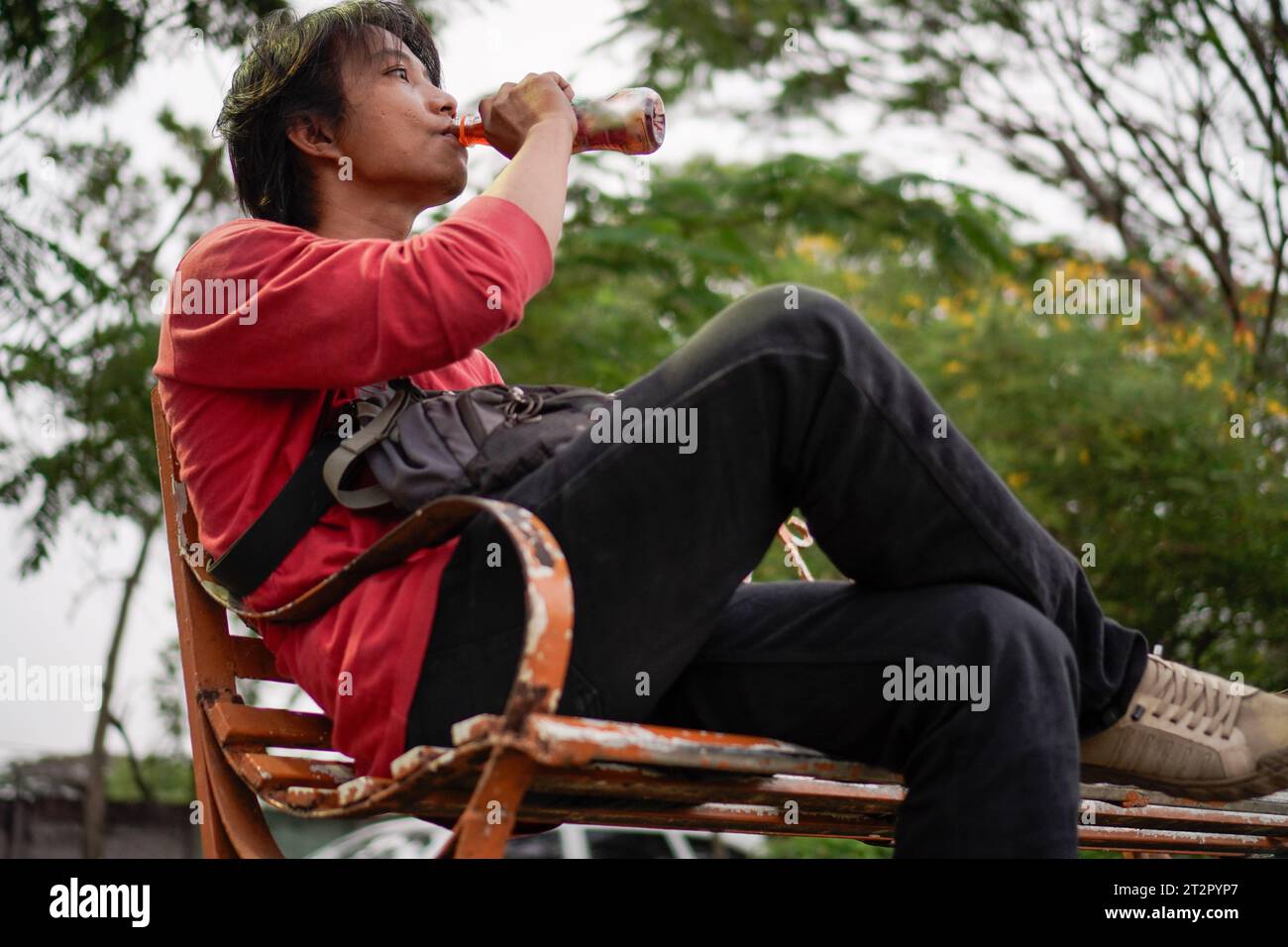 Javanese man in red shirt, sitting on a bench, plant background with a low angle view facing the sky. Stock Photo