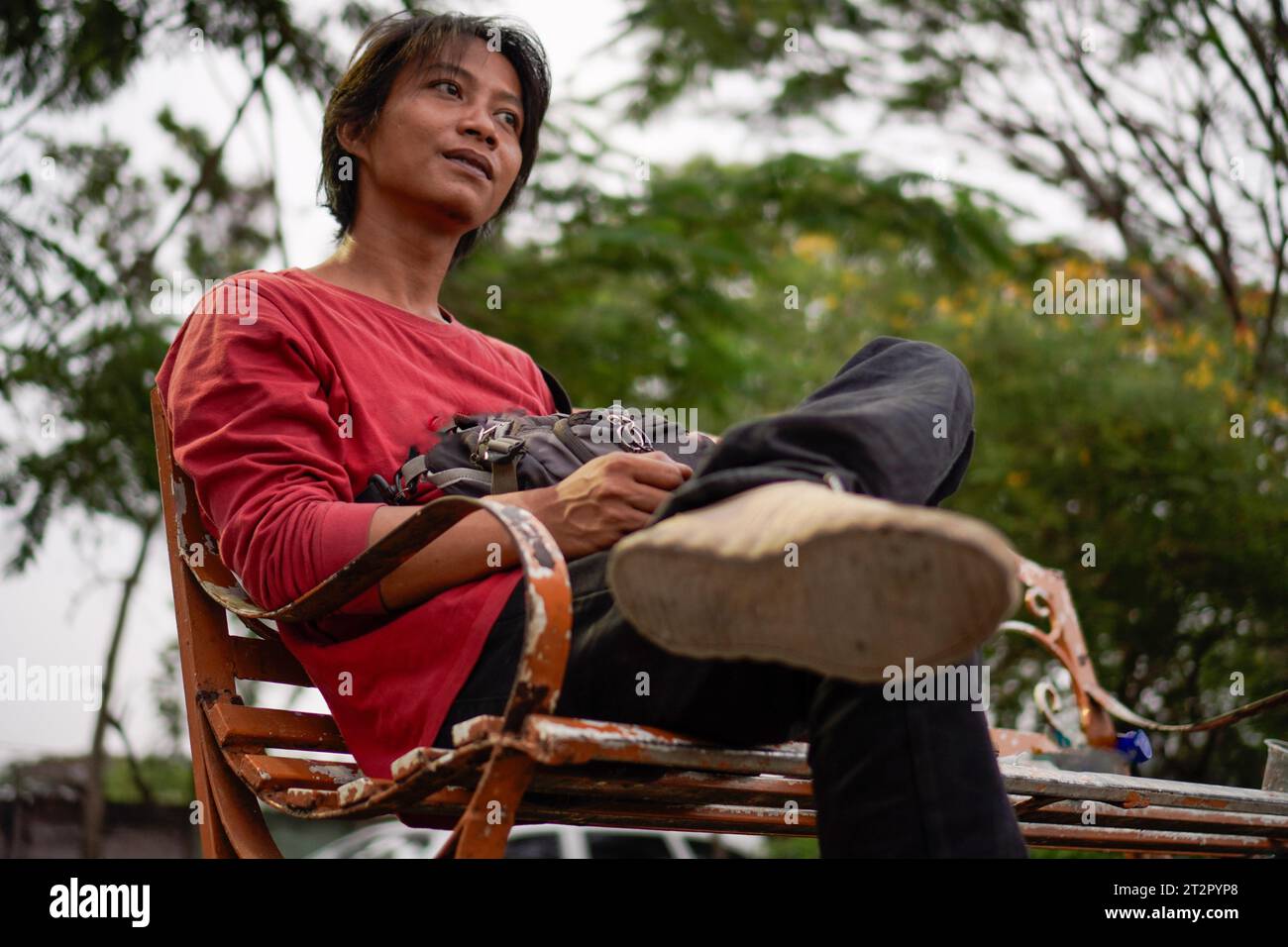 Javanese man in red shirt, sitting on a bench, plant background with a low angle view facing the sky. Stock Photo