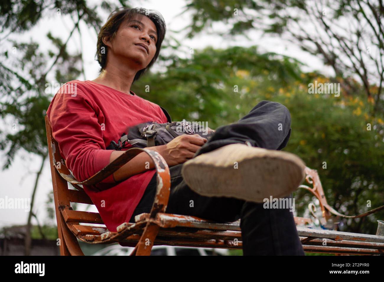 Javanese man in red shirt, sitting on a bench, plant background with a low angle view facing the sky. Stock Photo