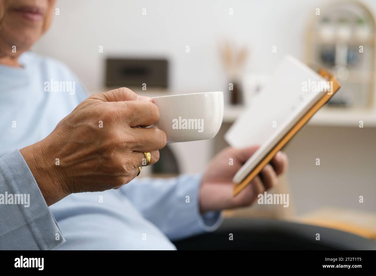 elderly man in eyeglasses sitting at home on the sofa reading a book and holding a cup of tea or coffee in his hand. Stock Photo
