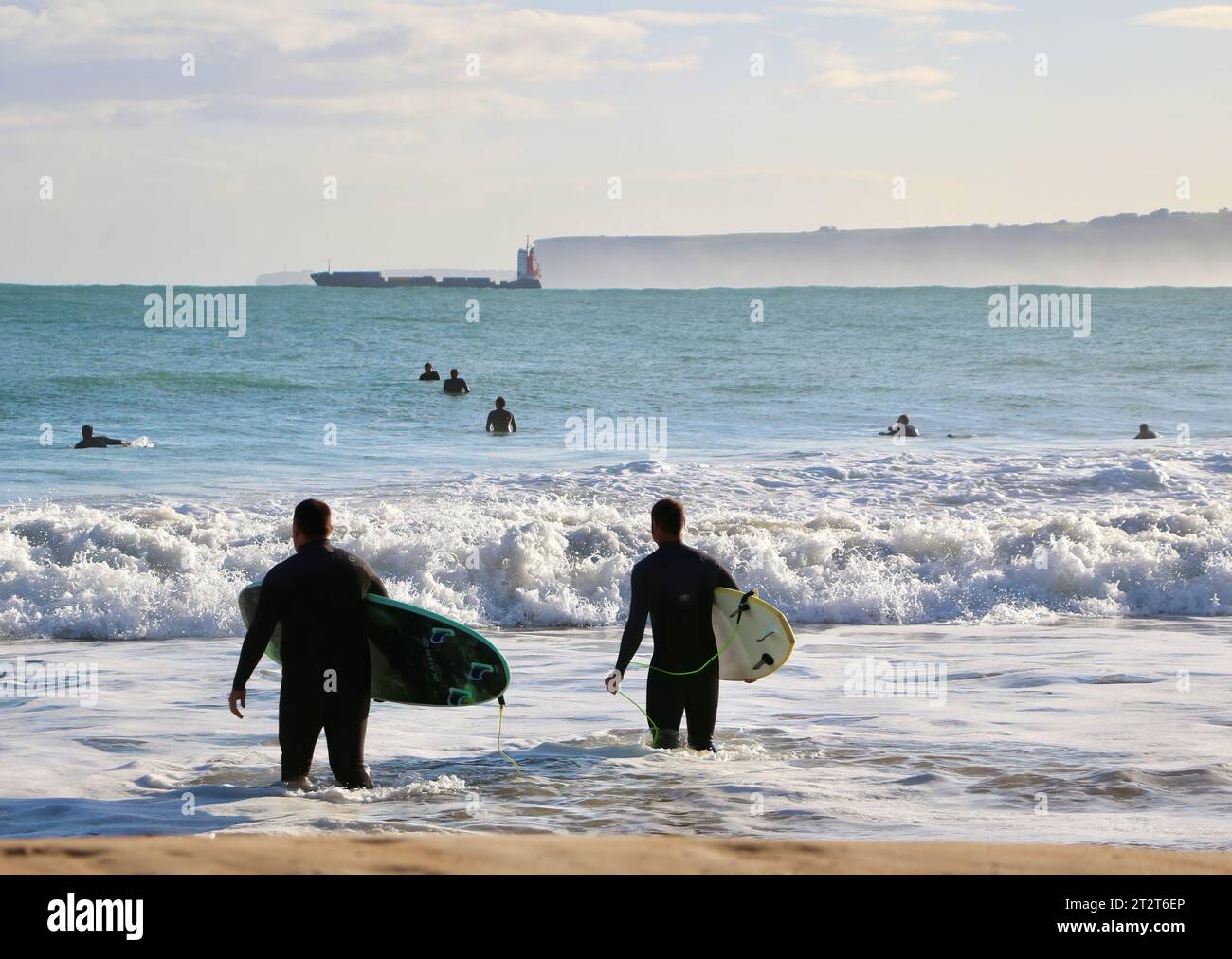 Surfers walking into the sea with a distant cargo ship leaving the port of Santander on a sunny October morning Sardinero Santander Cantabria Spain Stock Photo