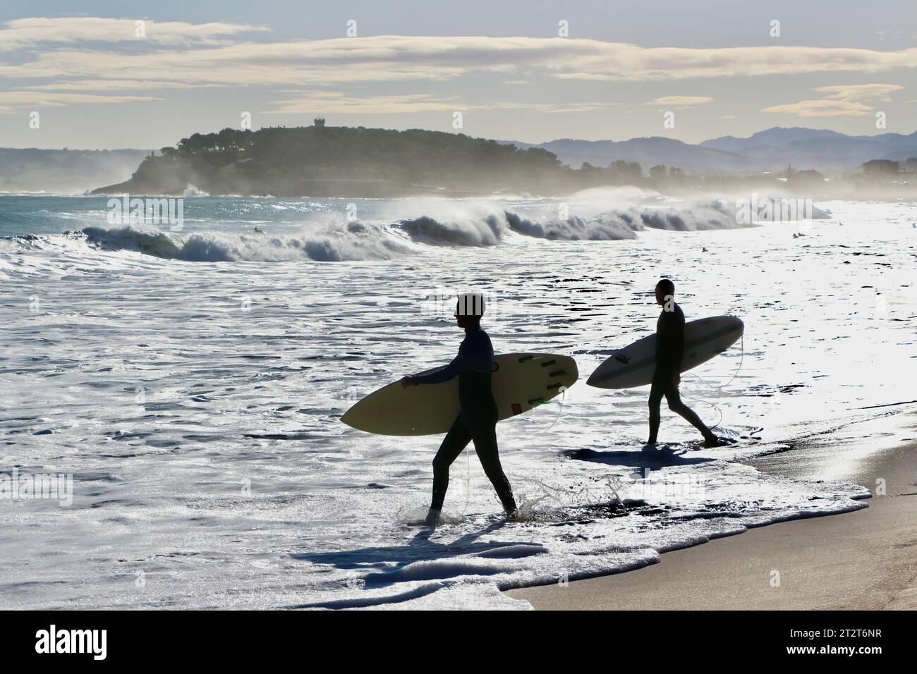 Autumn landscape with two surfers walking into the surf carrying surfboards backlit with the Magdalena Peninsular Sardinero Santander Cantabria Spain Stock Photo