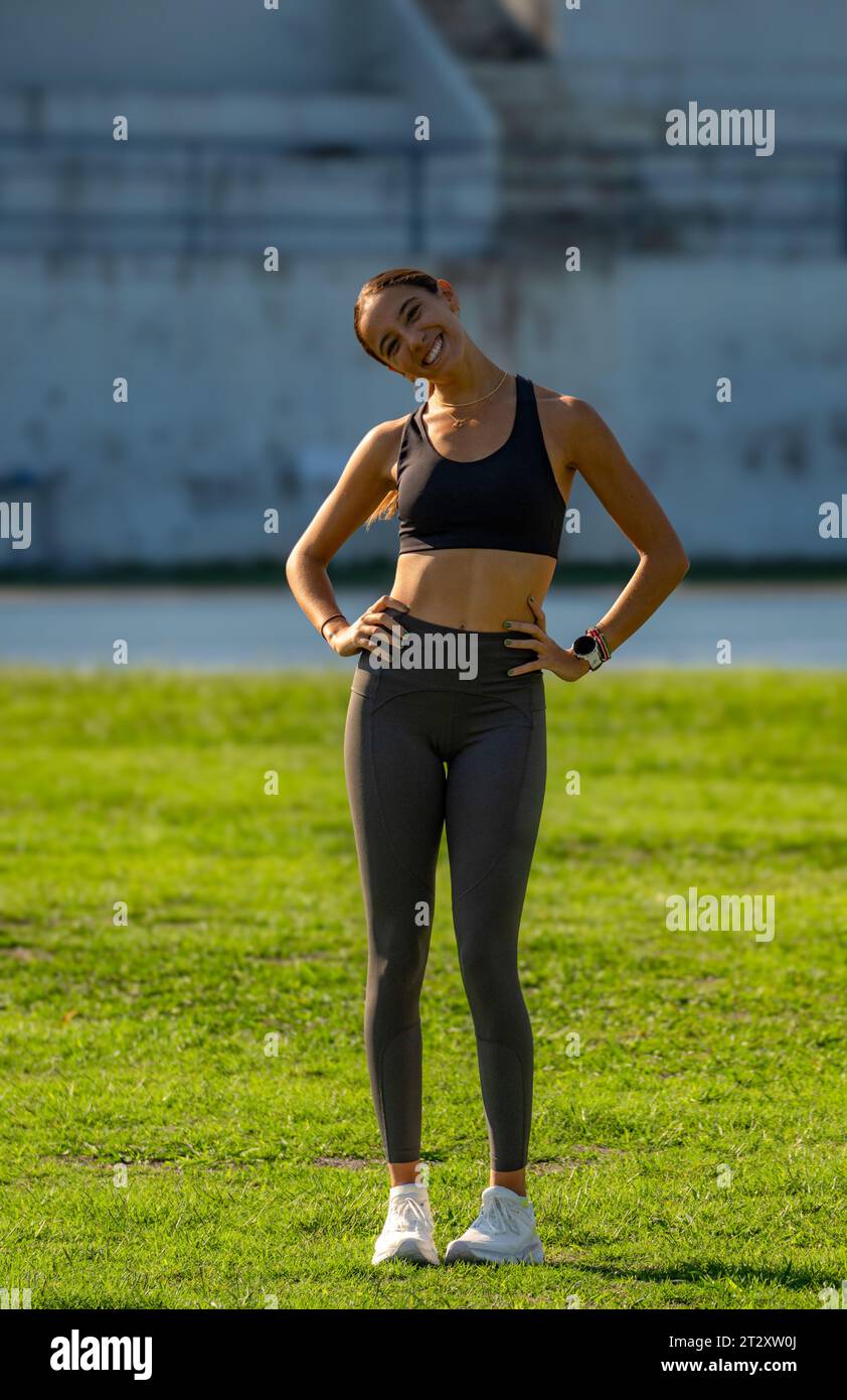 Beautiful young slim and tanned runner girl, dressed in tight sportswear, smiling funny with her head tilted and arms on her hips looking at the camer Stock Photo