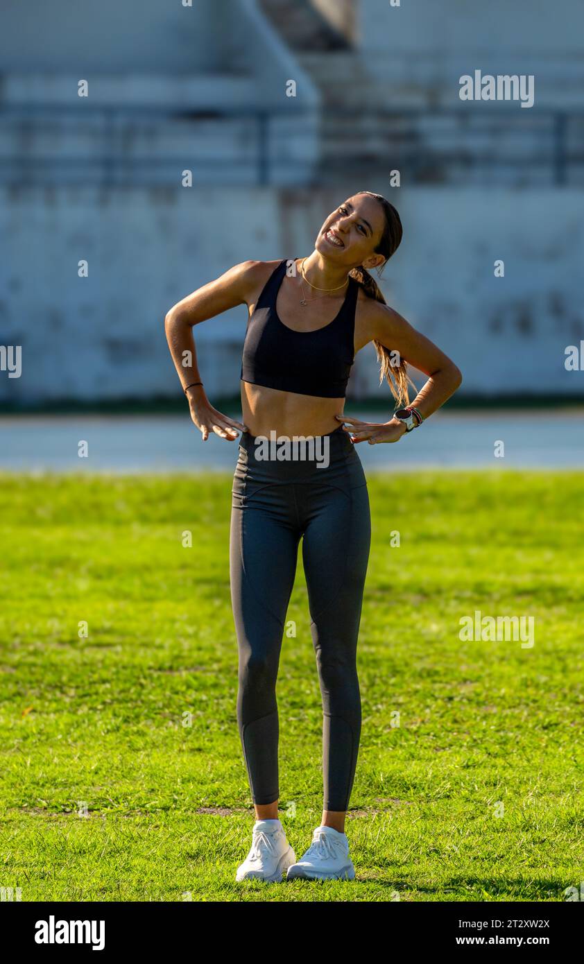 Beautiful slim and tanned young CrossFit girl, dressed in tight sportswear, smiling funny with her head tilted and arms on her hips looking at the cam Stock Photo