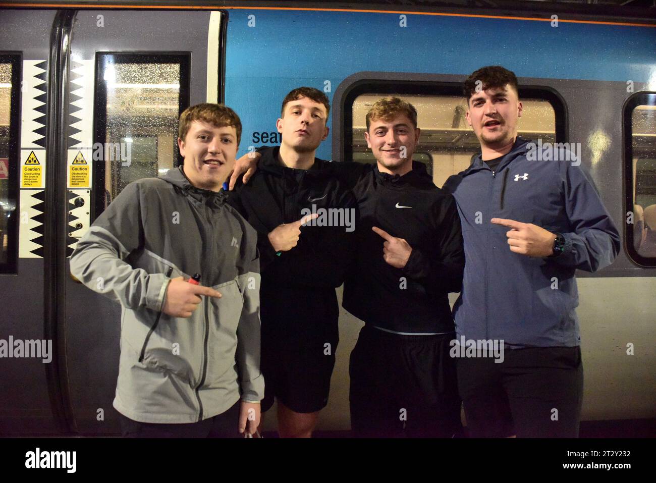 Four handsome young men wearing trendy sportswear pose at Liverpool Lime Street Railway Station where they are about to catch a train to Manchester, UK, at the height of Storm Babet, to go clubbing at the Warehouse Party at Mayfield. The finger hand gesture, I am told, translates as 'lemon' meaning cool, and is used increasingly by young people of this age group in the UK to show they oppose violence, discrimination, and extreme behavior and just want an enjoyable night out with like-minded people. Stock Photo