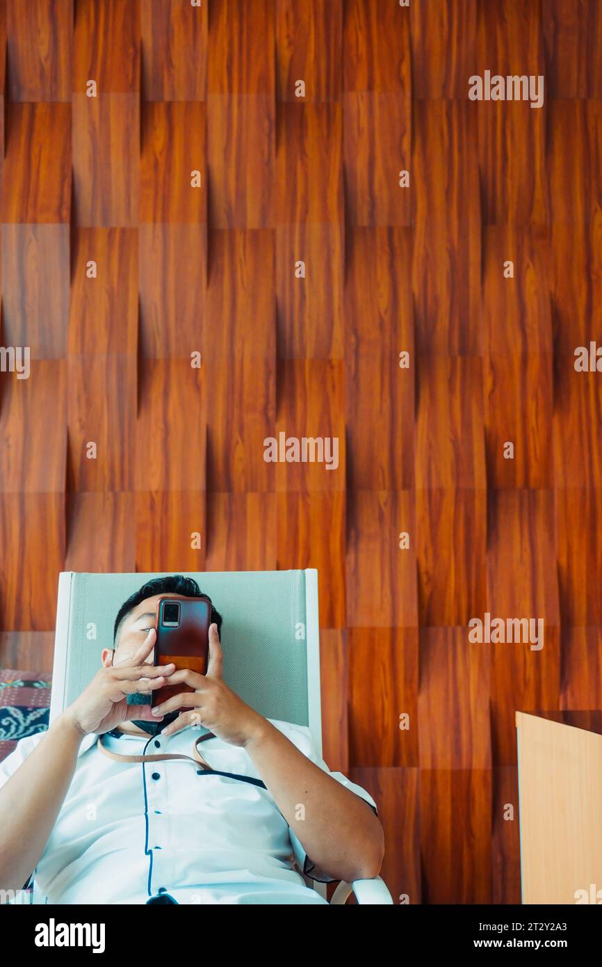 a man seated on a chair, engrossed in his mobile phone, set against a stylish wooden board background. The composition is carefully designed Stock Photo