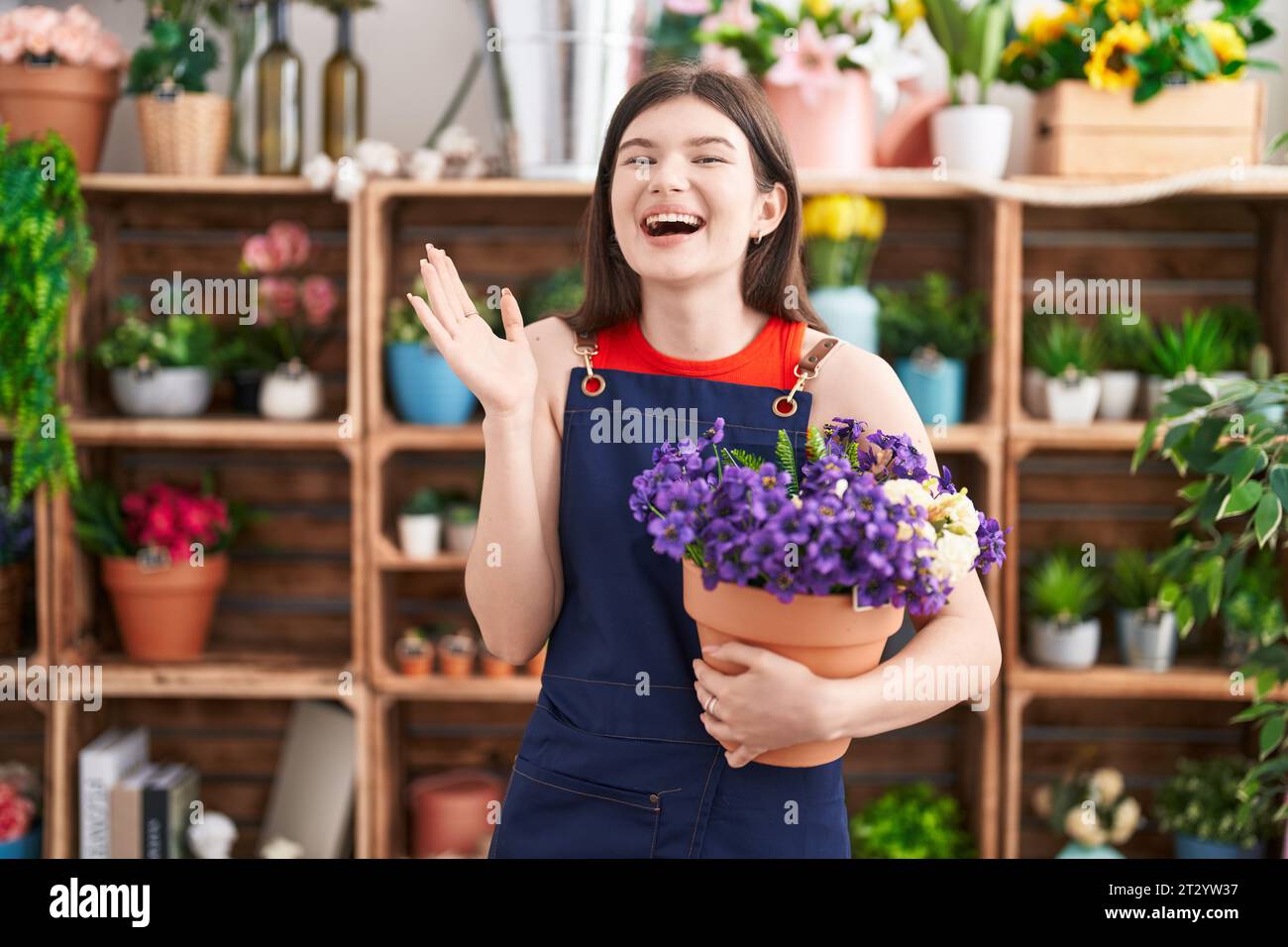 Young caucasian woman working at florist shop holding pot with flowers celebrating victory with happy smile and winner expression with raised hands Stock Photo