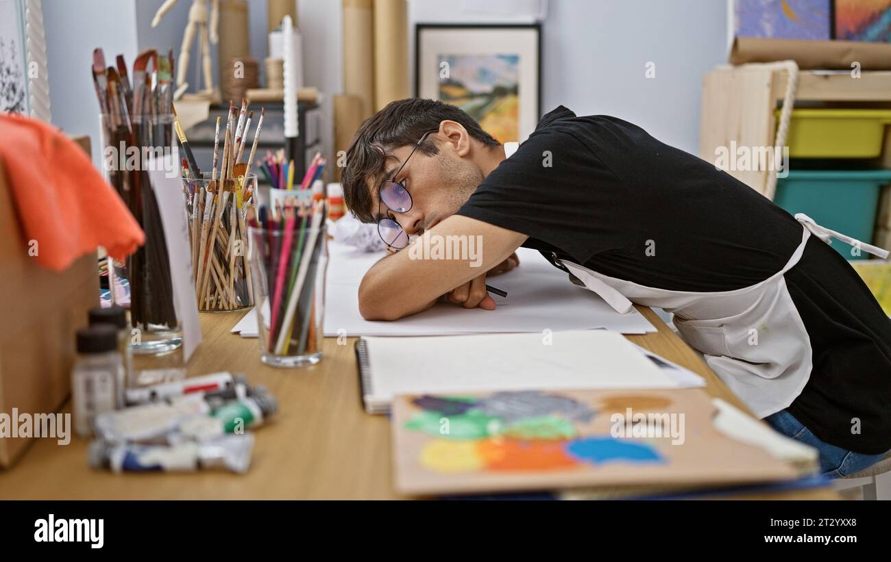 Exhausted young hispanic man, an art student, immersed in his drawing, leans on studio table, struggling with creativity, surrounded by paints, brushe Stock Photo