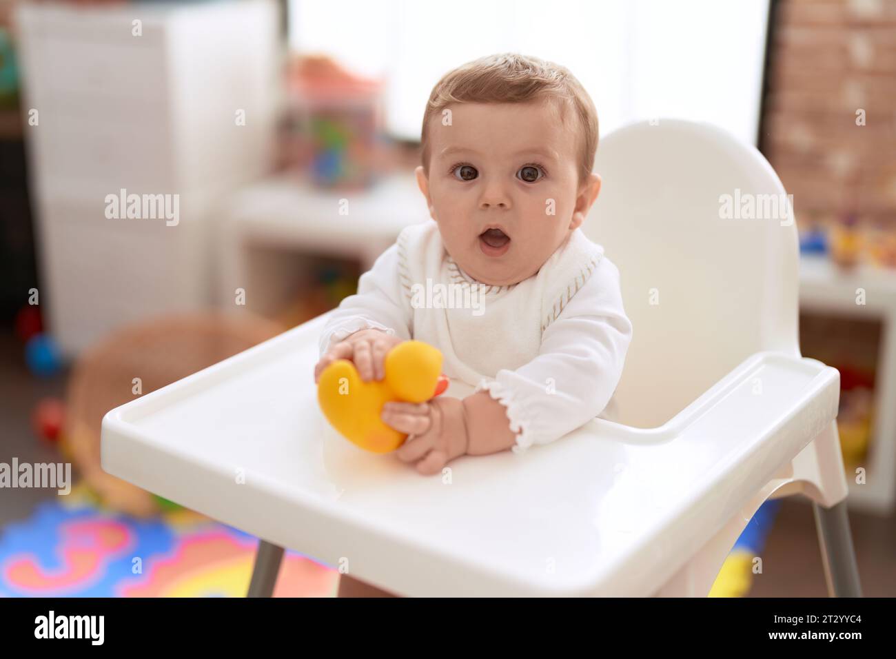 Adorable toddler sitting on baby highchair holding rubber duck toy at kindergarten Stock Photo