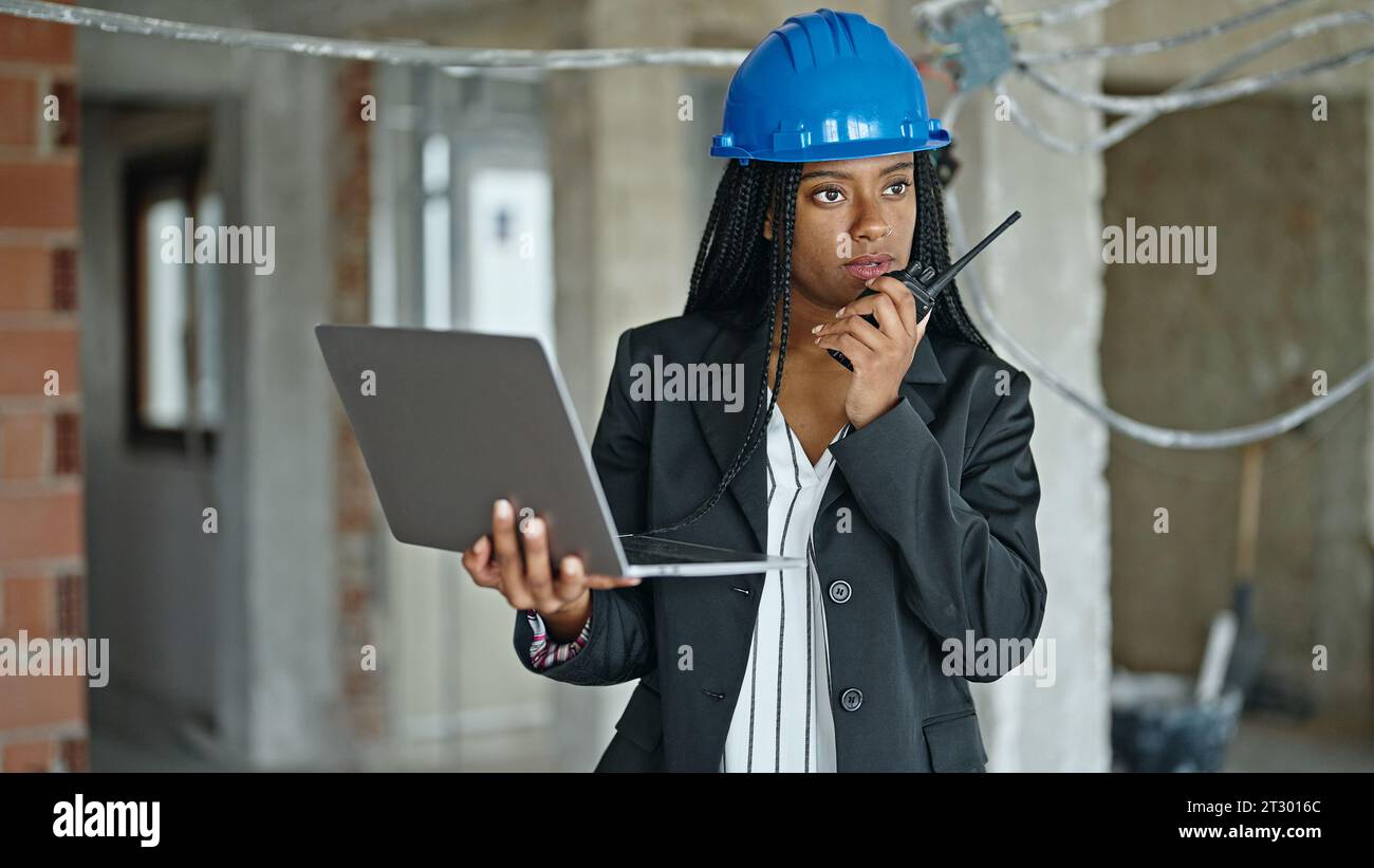 African american woman architect using laptop talking on walkie talkie at construction site Stock Photo