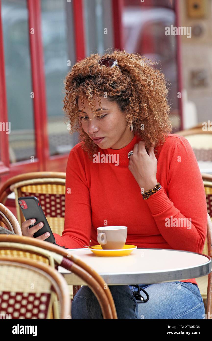 CURLY HAIR WOMAN IN RED SWEATER CHECKING HER PHONE ON A CHAIR OUTDOOR OF A COFFEE PLACE IN PARIS Stock Photo
