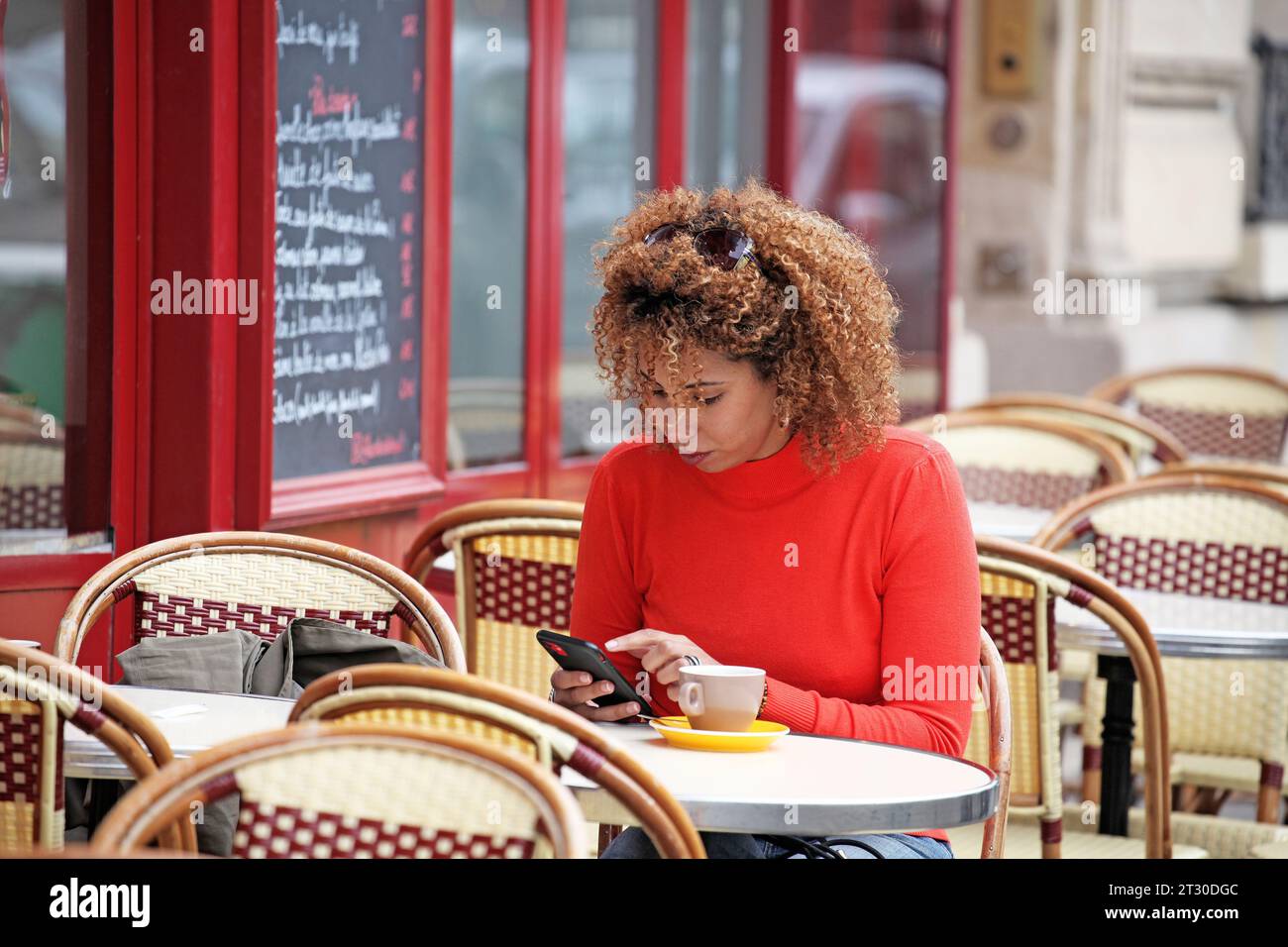 CURLY HAIR WOMAN IN RED SWEATER CHECKING HER PHONE ON A CHAIR OUTDOOR OF A COFFEE PLACE IN PARIS Stock Photo