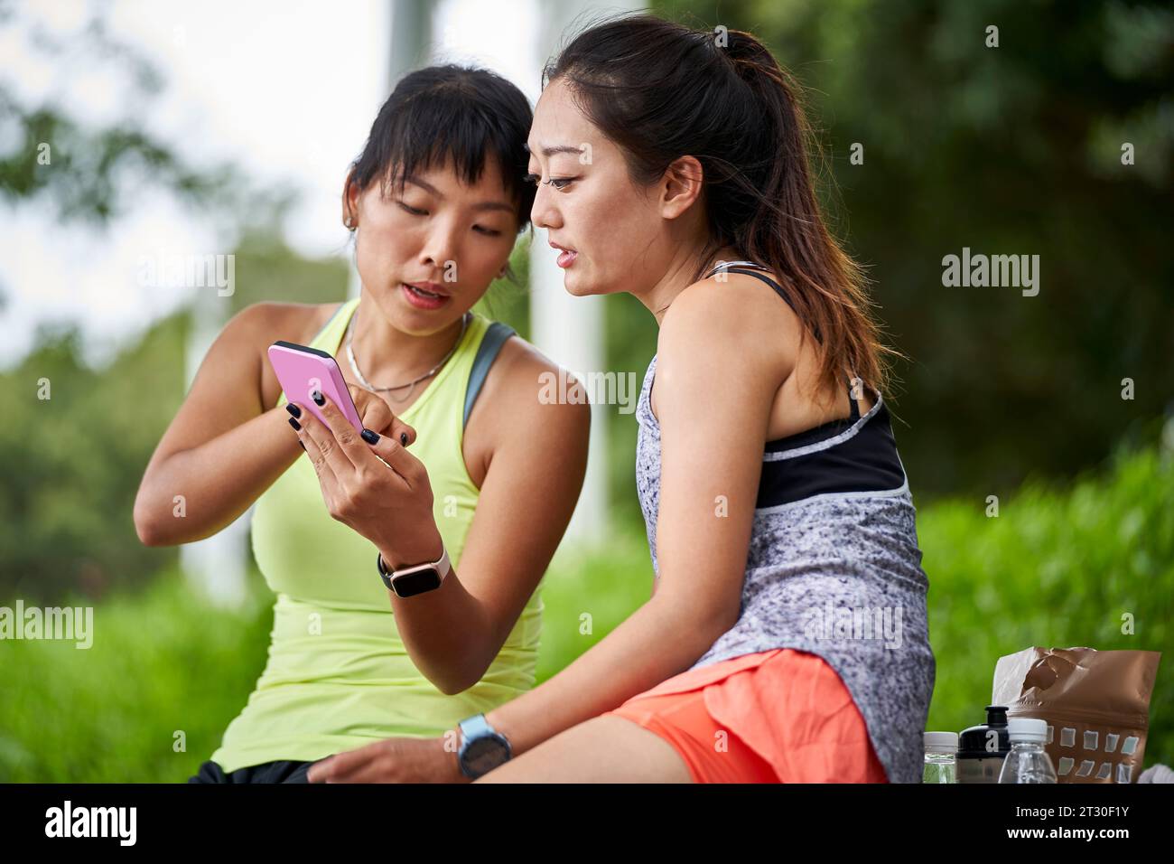 two young asian women female friends in sportswear relaxing chatting sharing cellphone photos outdoors Stock Photo