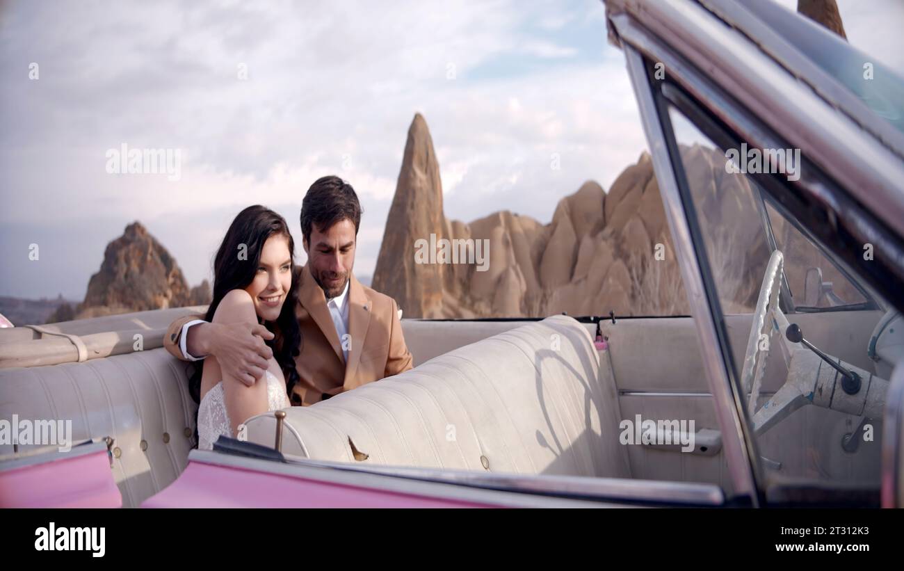 Happy bride and goom embracing while sitting in pink cabriolet car with stone formations behind. Action. Photosession outdoors. Stock Photo