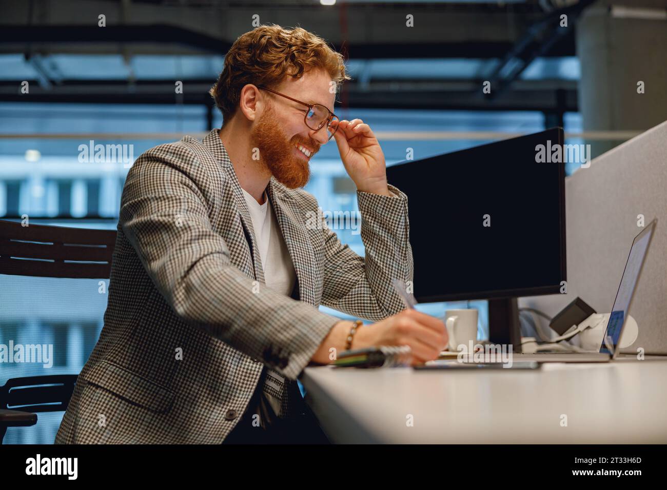 Smiling businessman working on laptop and making notes while sitting desk on office background Stock Photo