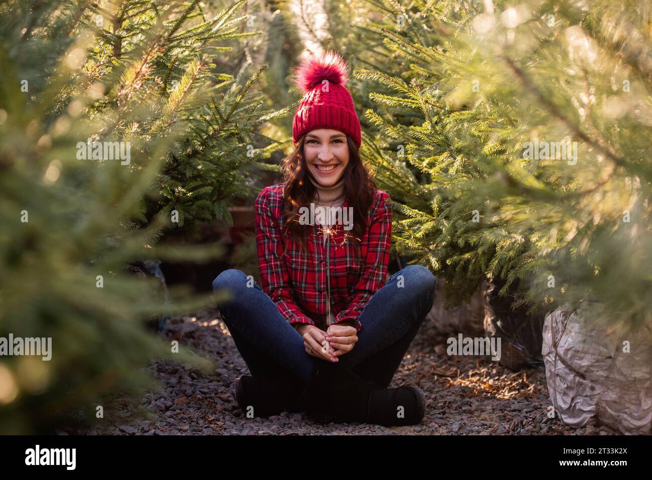 Young woman with sparkler in hand wearing red checkered shirt, knitted hat among green Christmas tree market. Curly-haired girl laughs and rejoices in Stock Photo