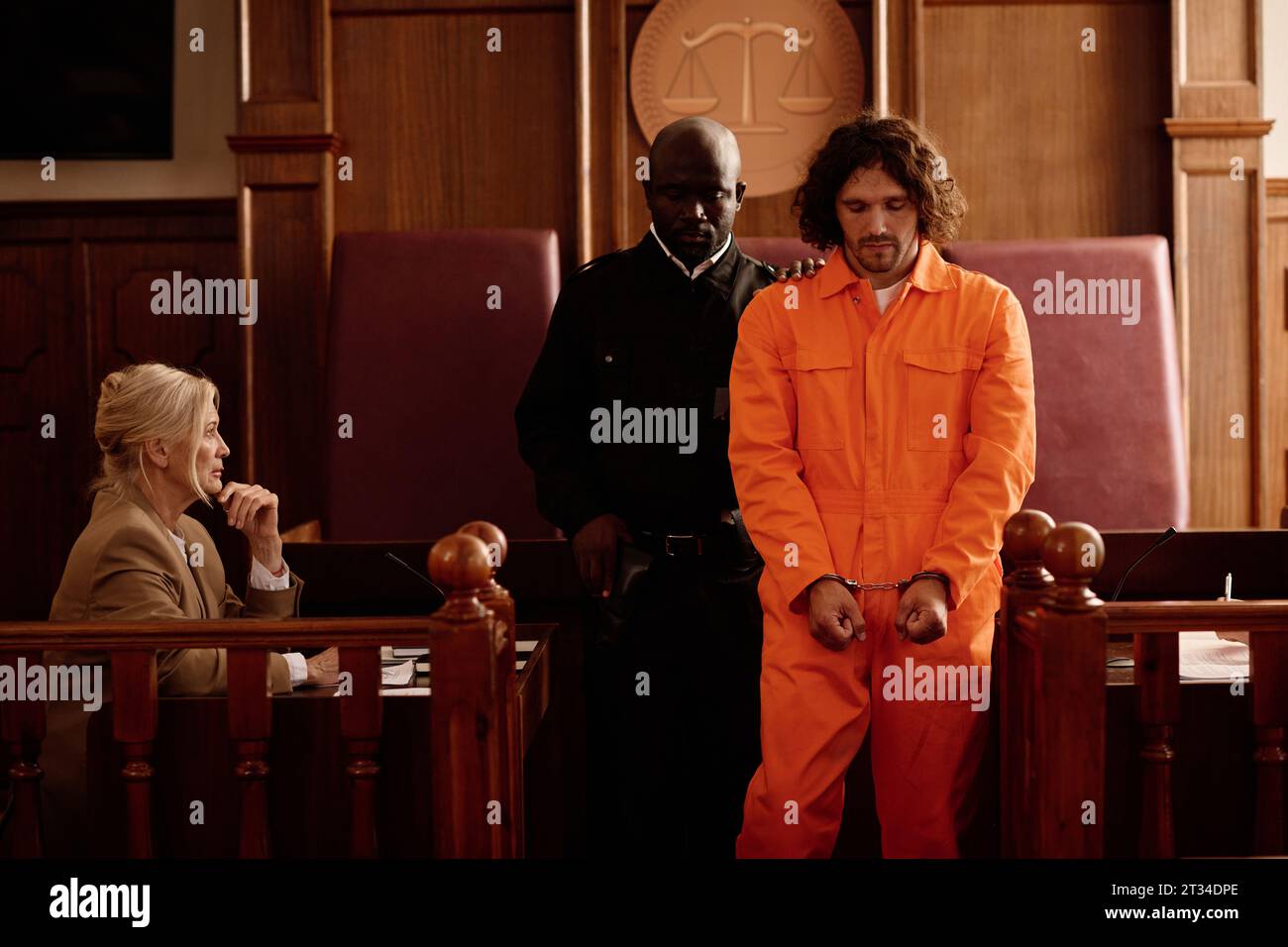 African American policeman standing next to young male prisoner in cuffs and orange jumpsuit while mature female witness testifying in court Stock Photo