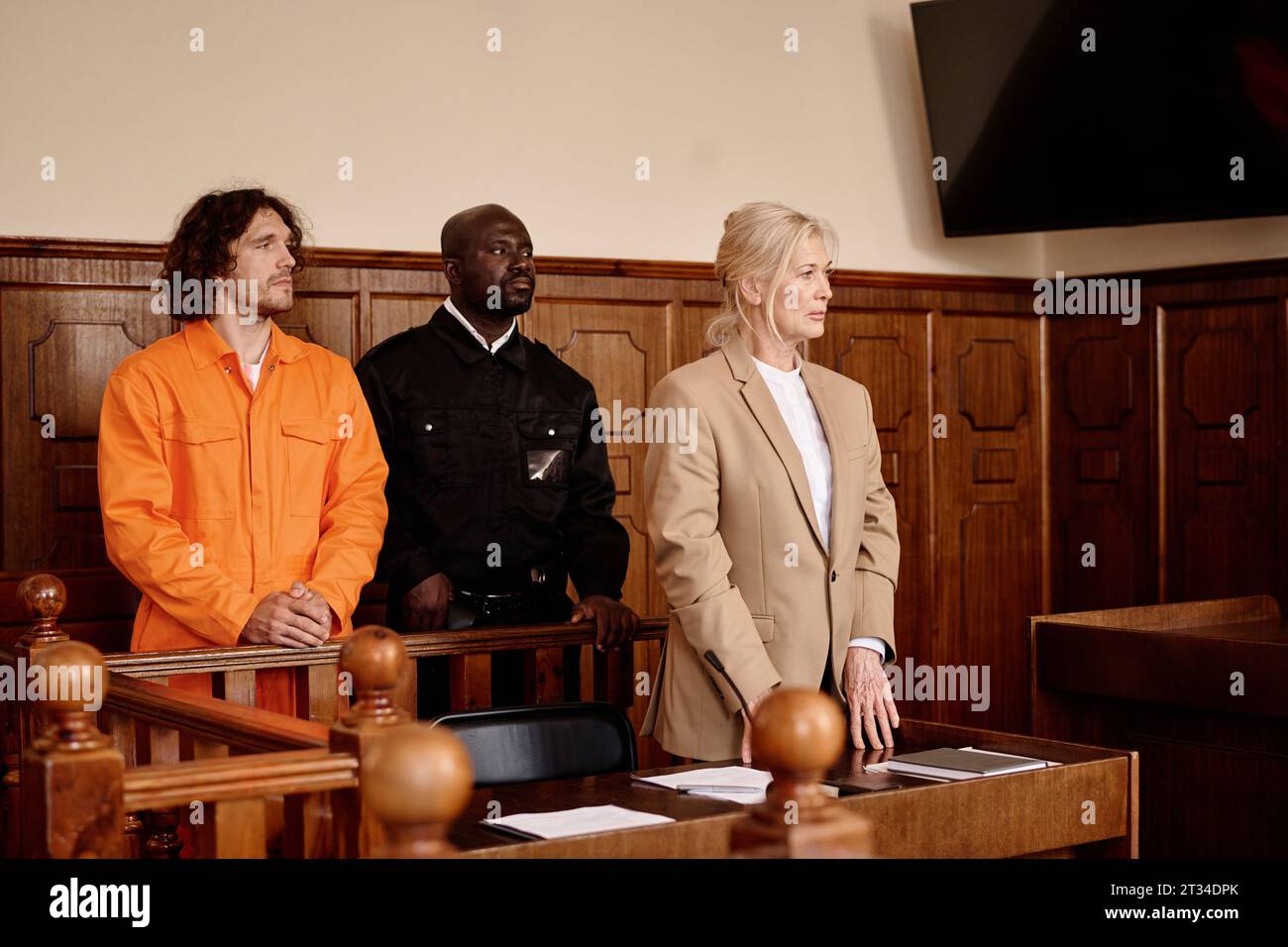 African American policeman standing between young male prisoner in cuffs and orange jumpsuit and mature female attorney during trial session Stock Photo