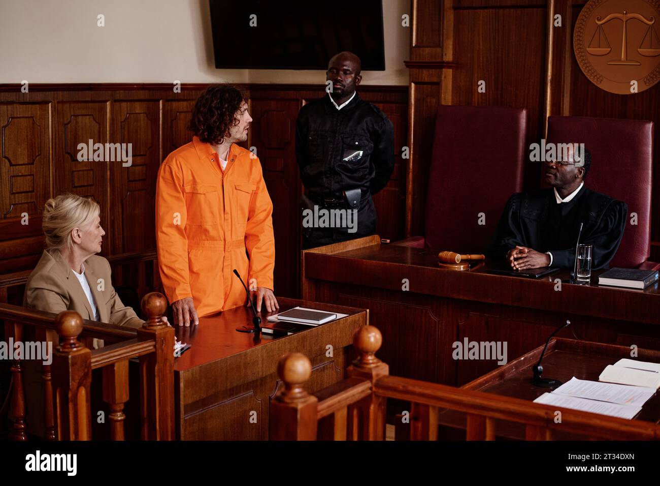 Young male suspect in orange jumpsuit standing next to his attorney and looking at mature African American impartial judge during trial session Stock Photo