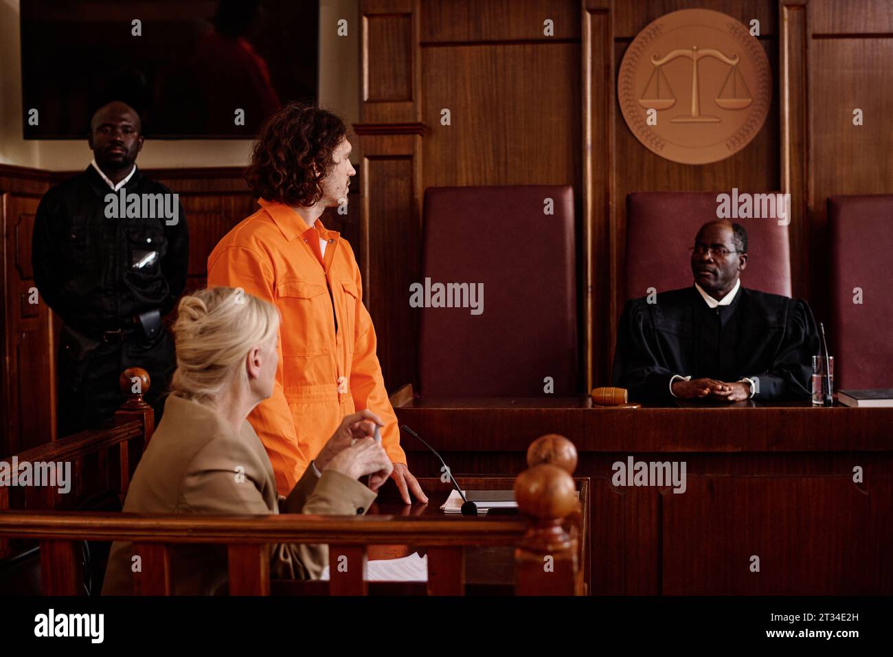 Side view of suspect in orange jumpsuit and his attorney looking at mature African American impartial judge while prisoner speaking to him Stock Photo