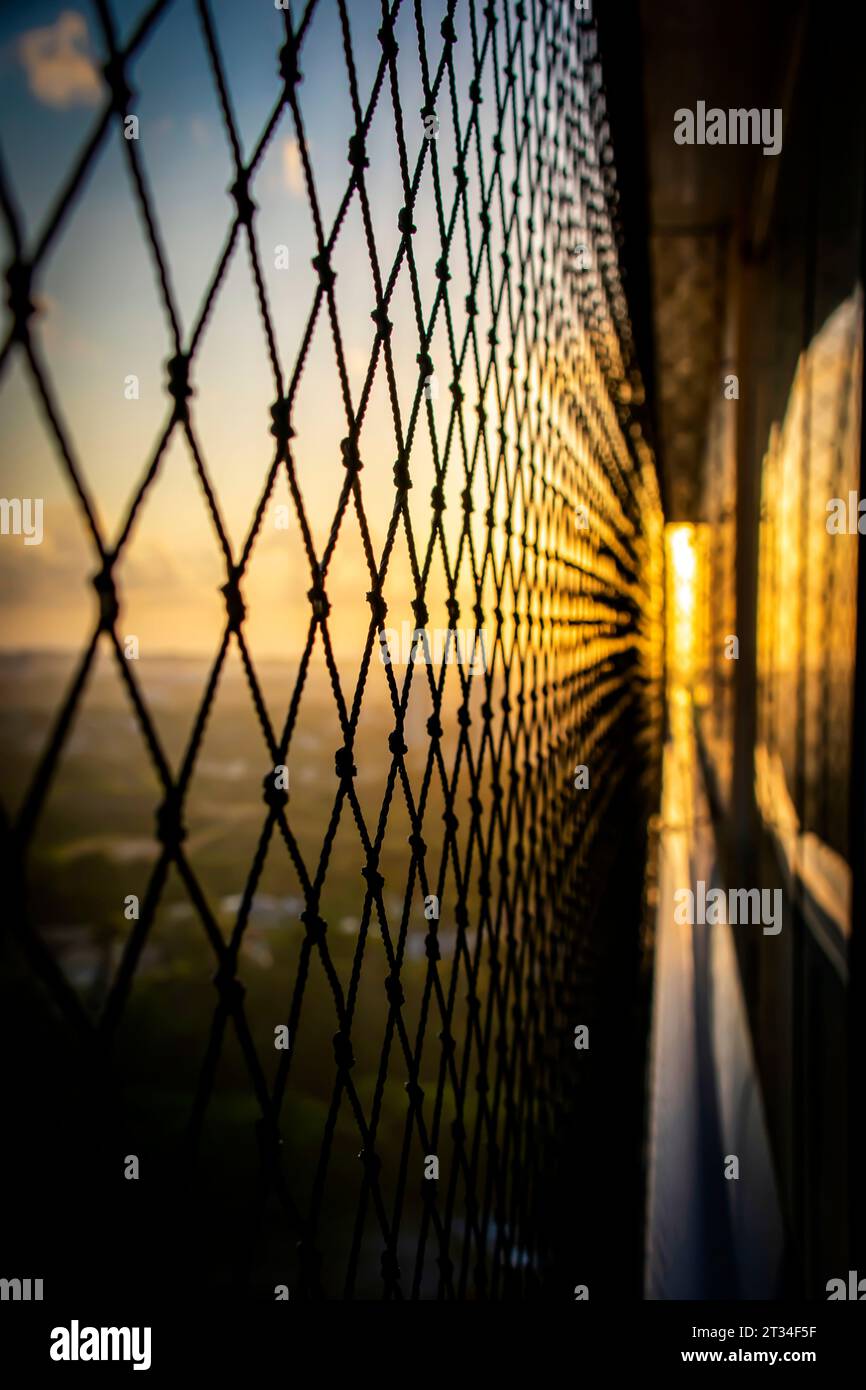 Sunrise glow streaming through a safety net on a balcony, casting intricate shadows and soft golden light over the view below. Stock Photo
