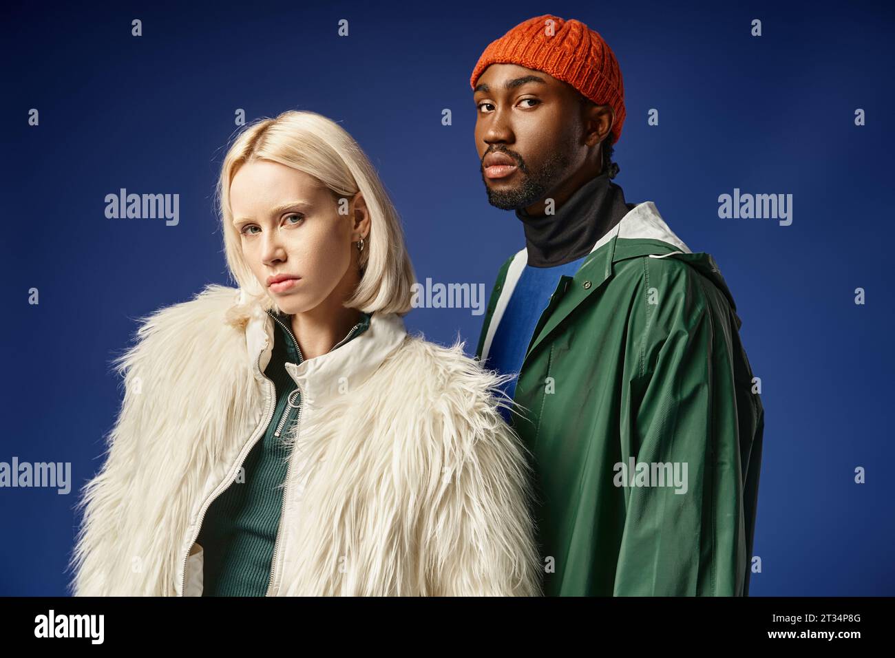 blonde woman posing with african american man in winter attire, trendy models on blue backdrop Stock Photo