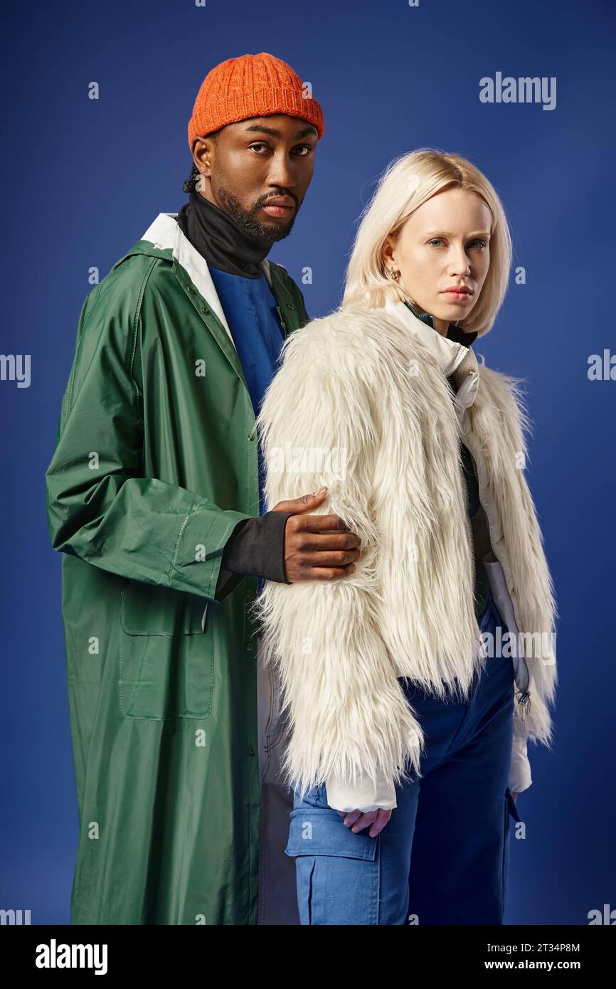 blonde woman posing with african american man in winter attire, stylish models on blue backdrop Stock Photo
