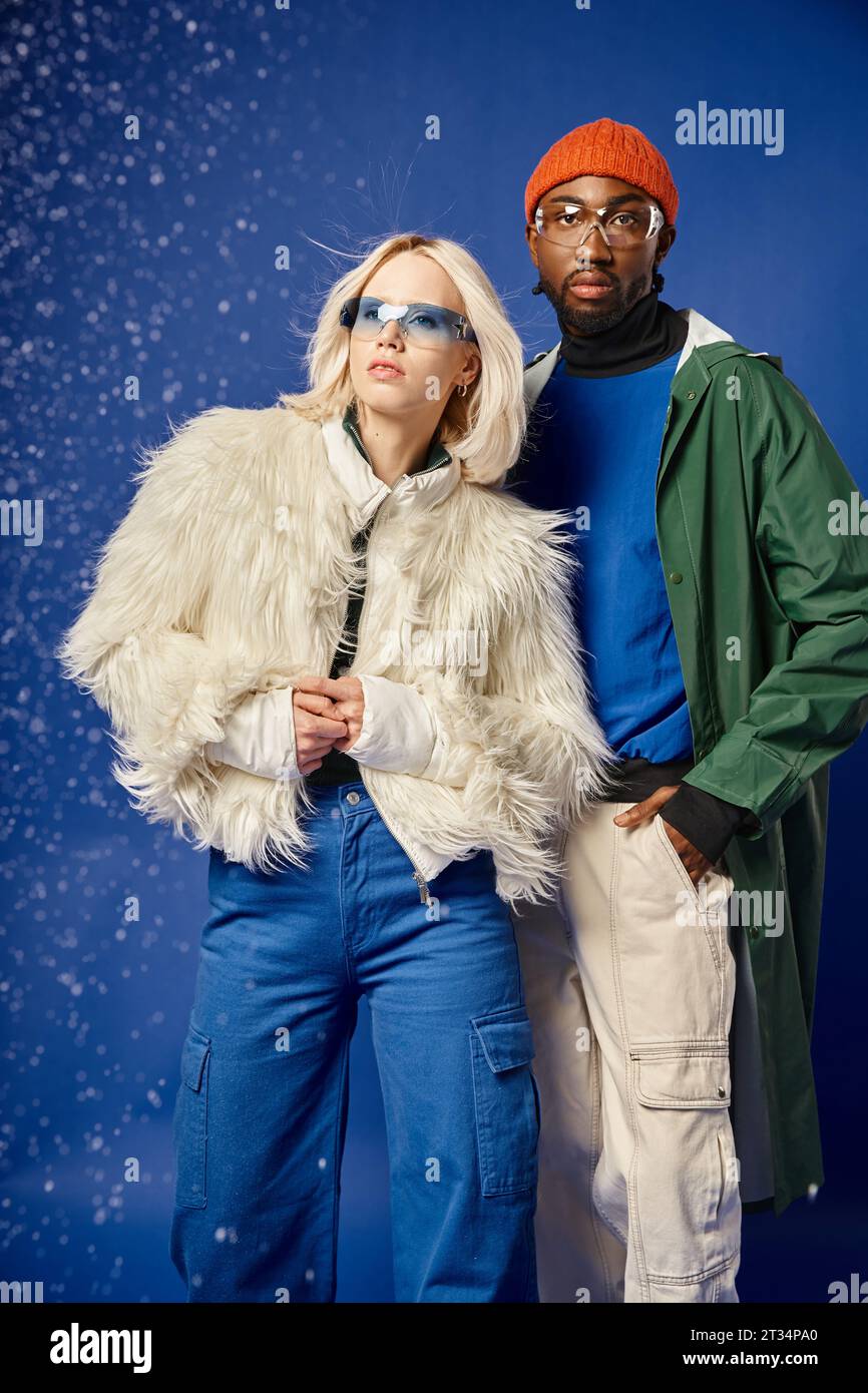 african american man and blonde woman posing in winter attire under falling snow on blue backdrop Stock Photo