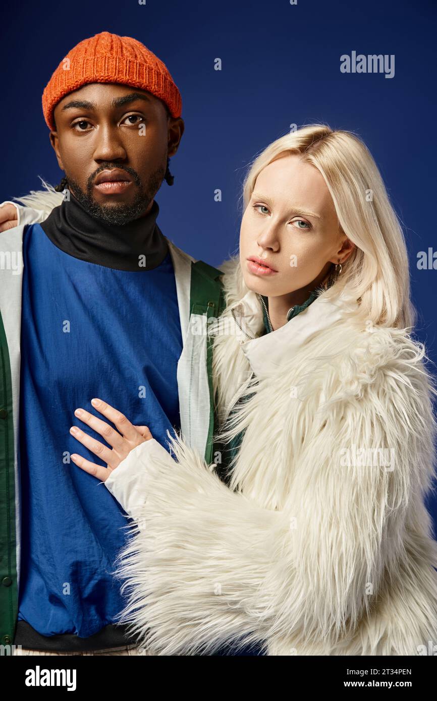 blonde woman embracing african american man in winter attire of blue backdrop, multiethnic couple Stock Photo