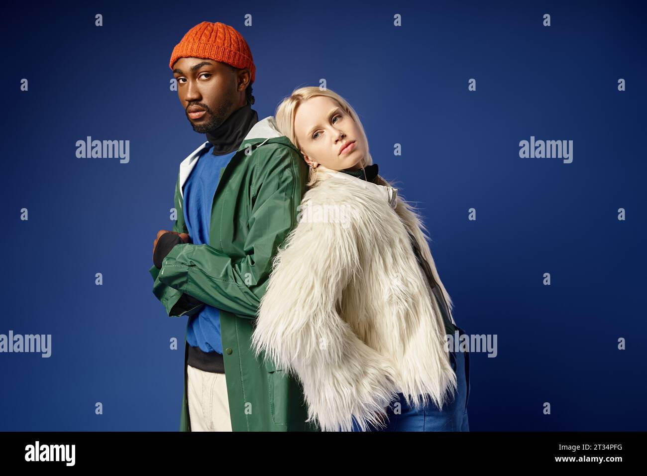blonde woman leaning on african american man in winter attire, diverse couple on blue backdrop Stock Photo