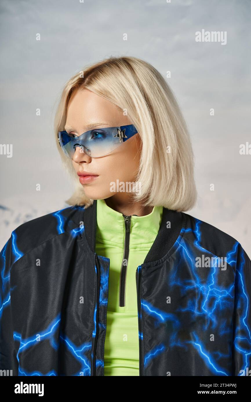 blonde young woman in blue sunglasses and stylish winter outfit looking away with snowy backdrop Stock Photo