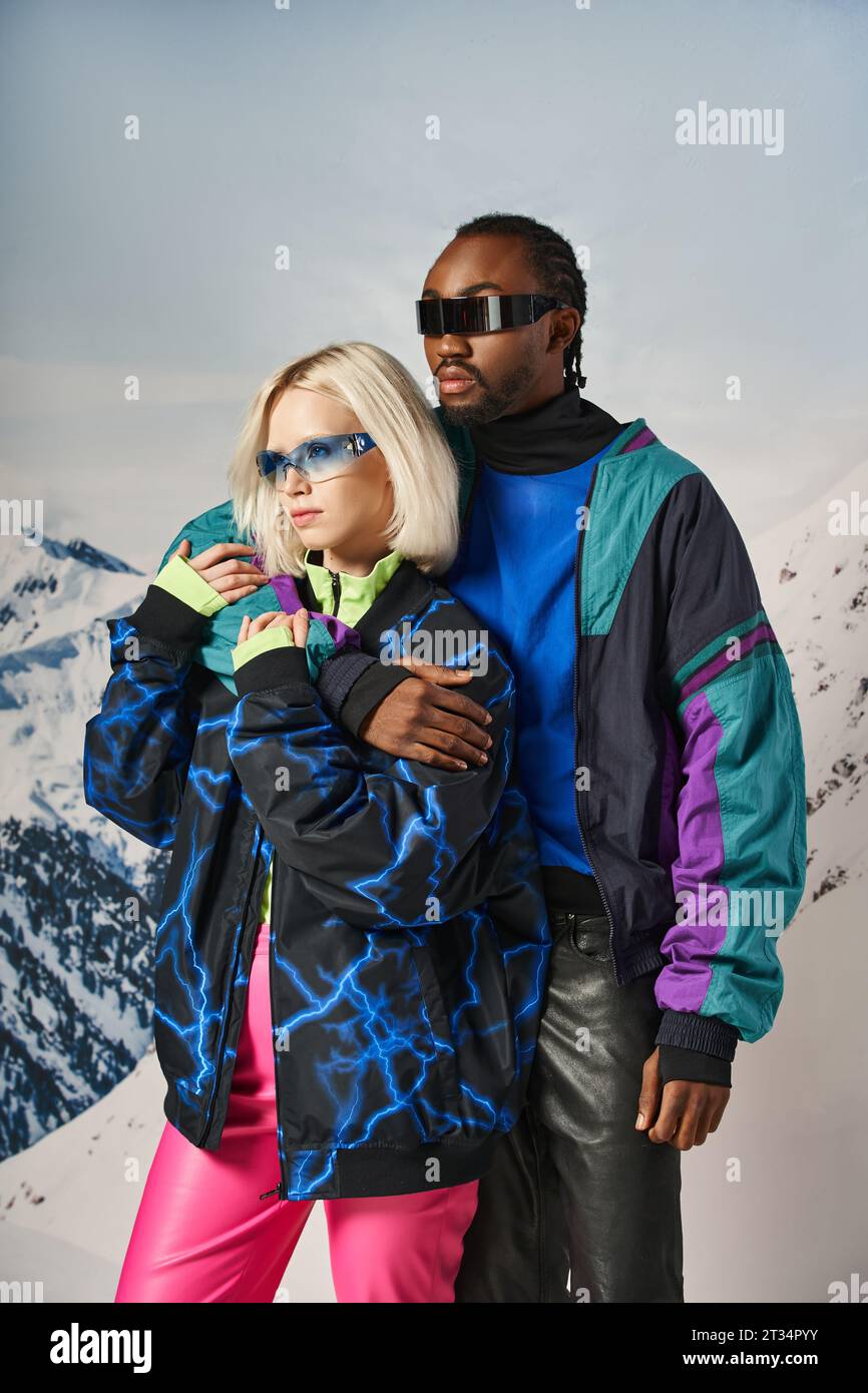 beautiful multiracial couple in vibrant bold outfits and stylish sunglasses hugging warmly, winter Stock Photo