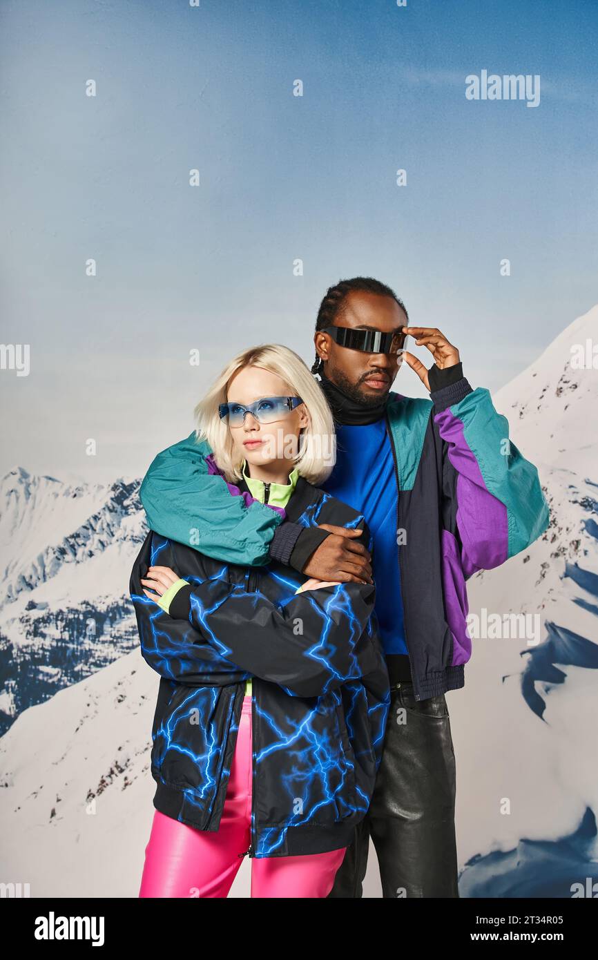 fashionable multiethnic couple in winter outfits with stylish glasses hugging, mountain backdrop Stock Photo