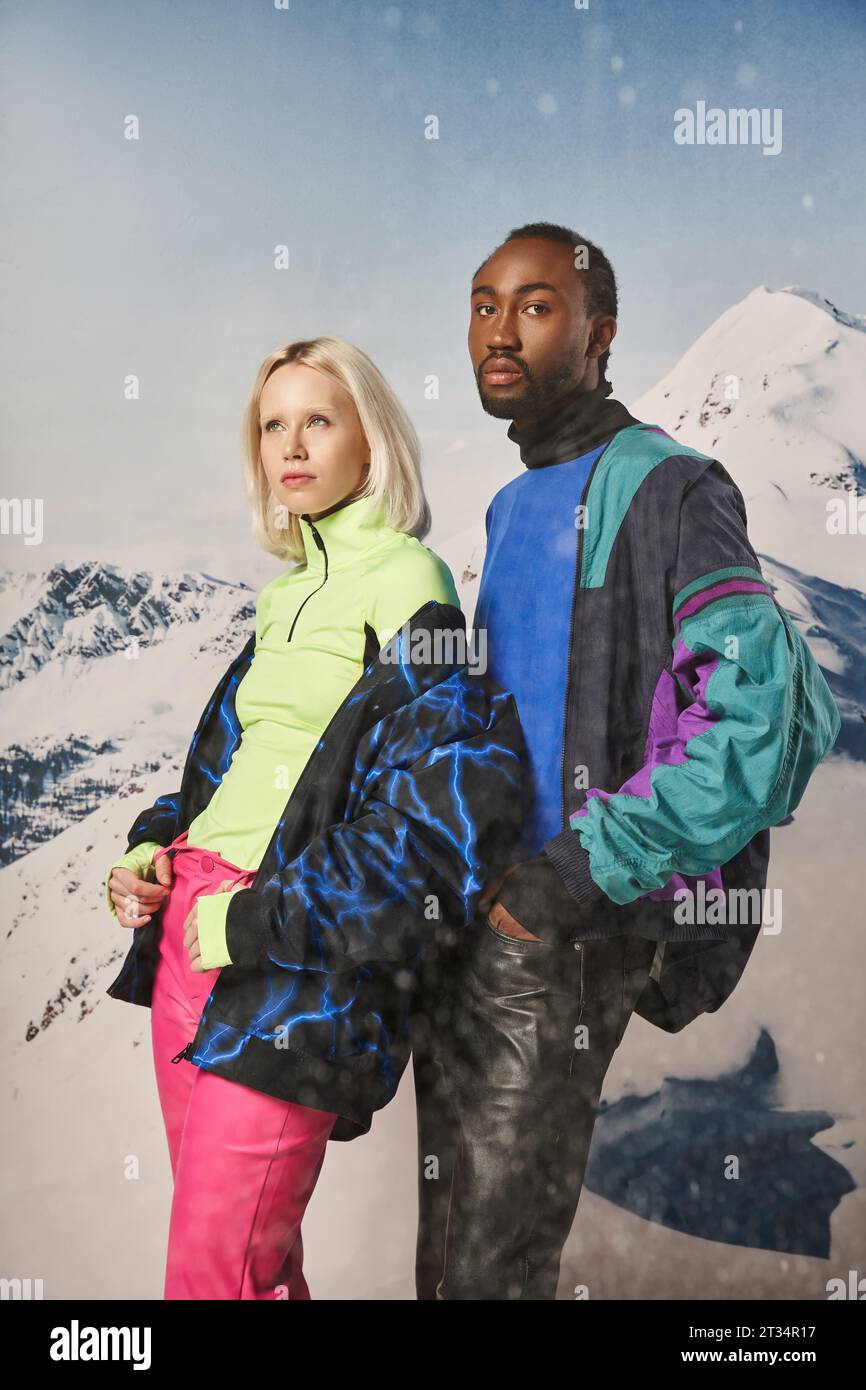 attractive diverse couple in stylish warm outfits with snowy mountain on backdrop, winter concept Stock Photo