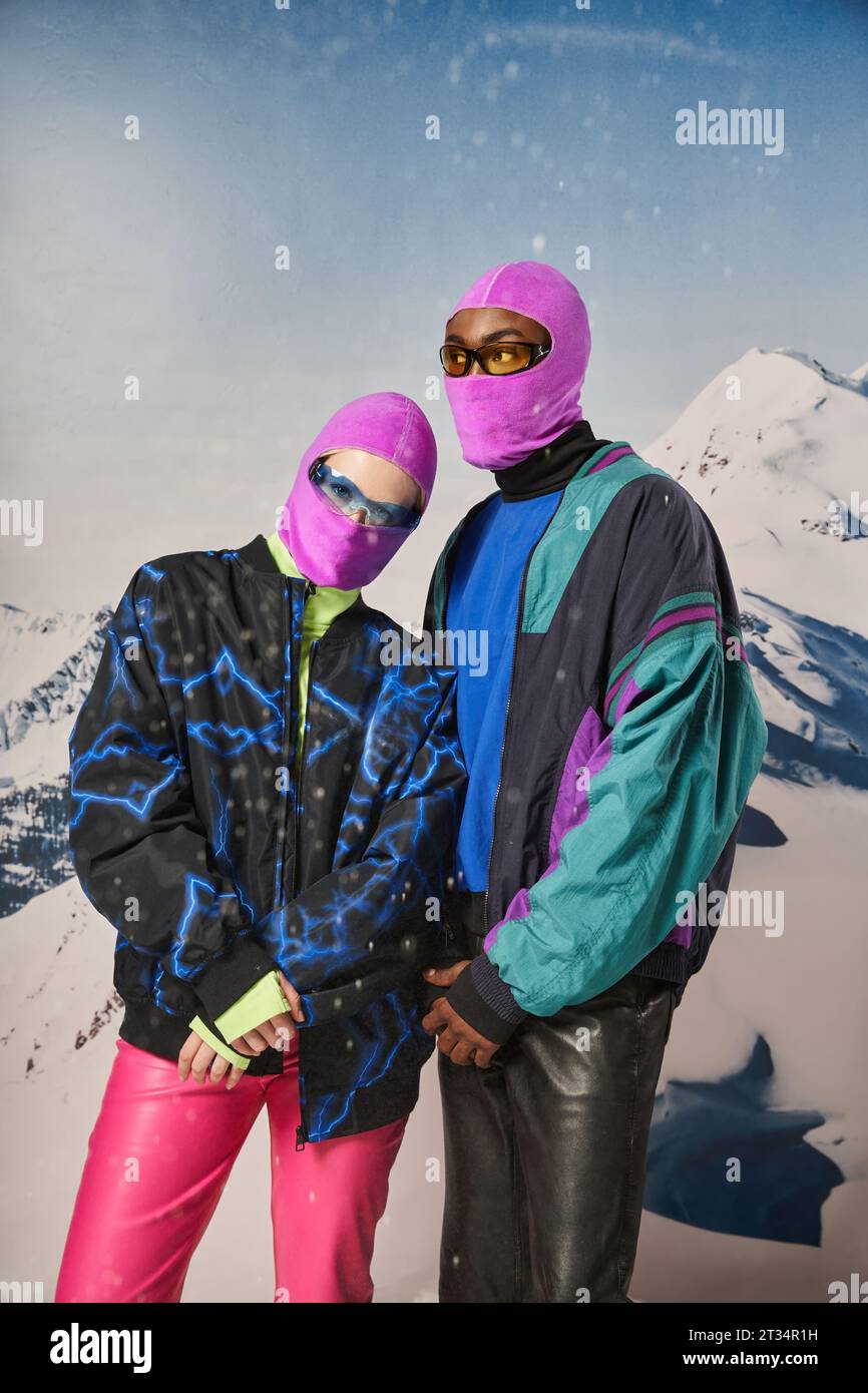 stylish young couple in warm winter outfits and balaclavas posing together on snowy backdrop Stock Photo