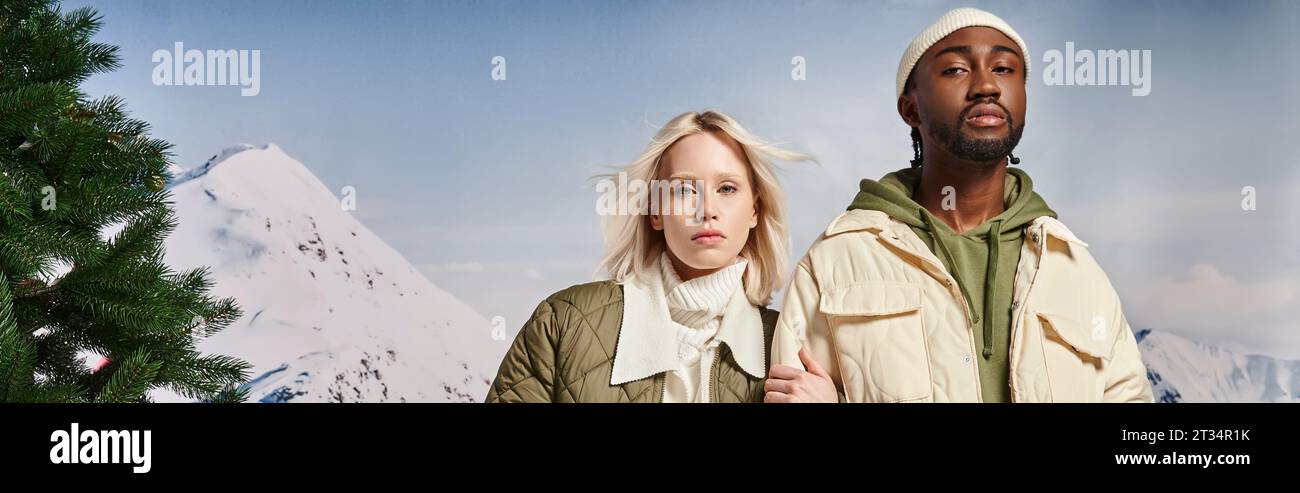 fashionable multiracial couple posing with hand on arm and looking at camera, winter concept, banner Stock Photo
