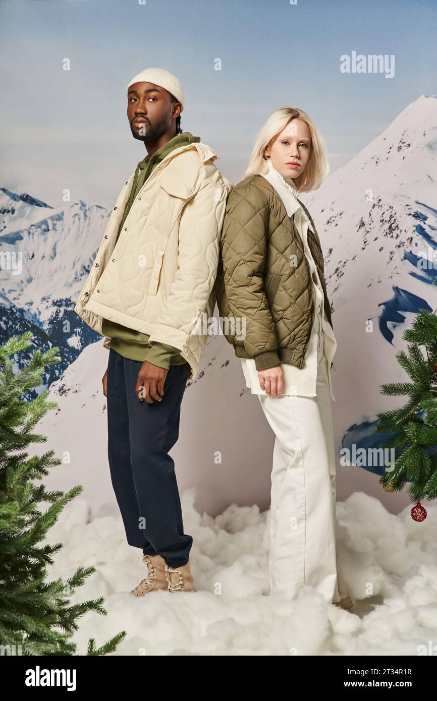 attractive young couple in warm outfits posing back to back with snowy background, winter concept Stock Photo