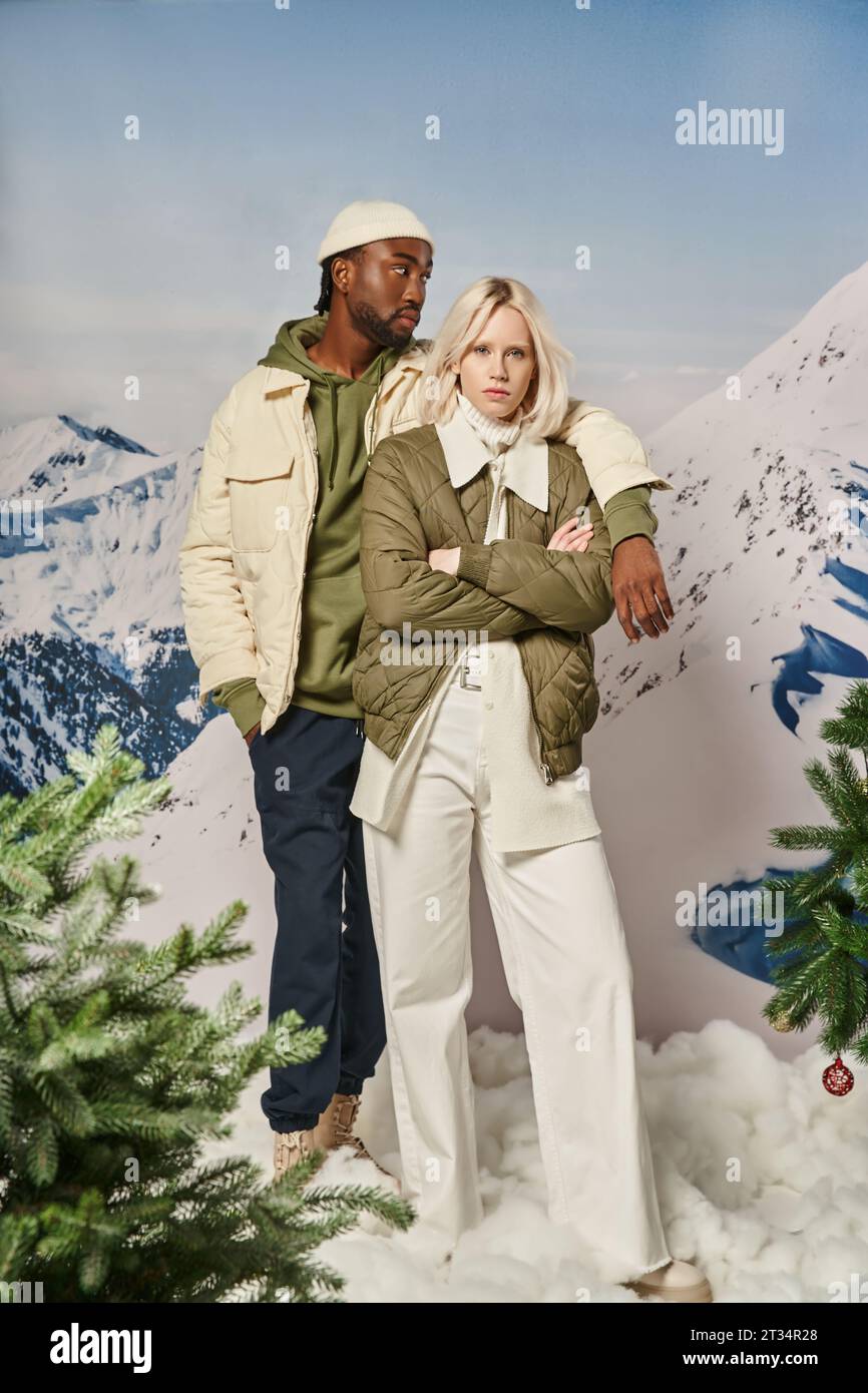 beautiful voguish couple in warm winter jackets with mountain backdrop posing with arm on shoulder Stock Photo