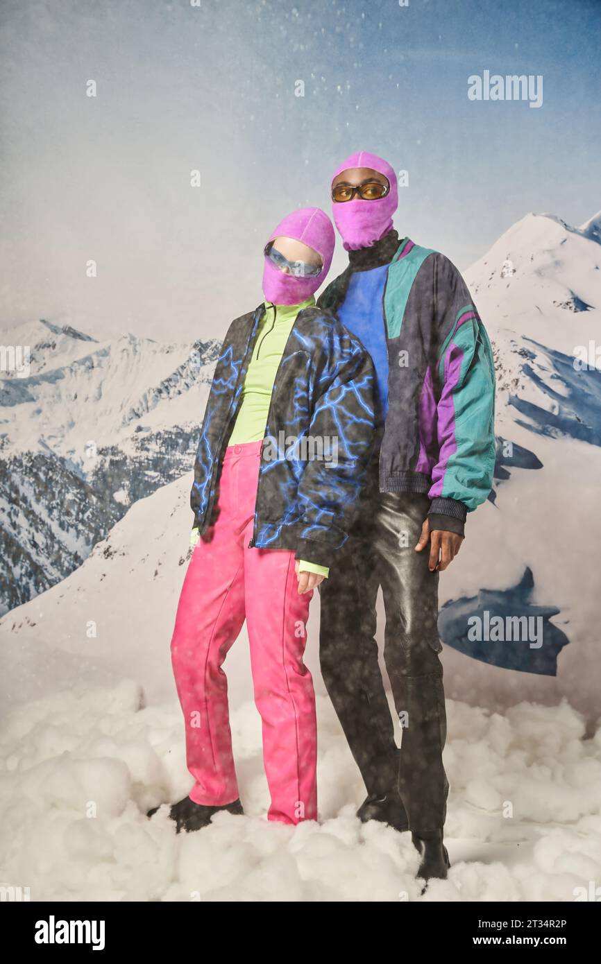 attractive diverse couple posing together in pink balaclavas with mountain backdrop, winter concept Stock Photo