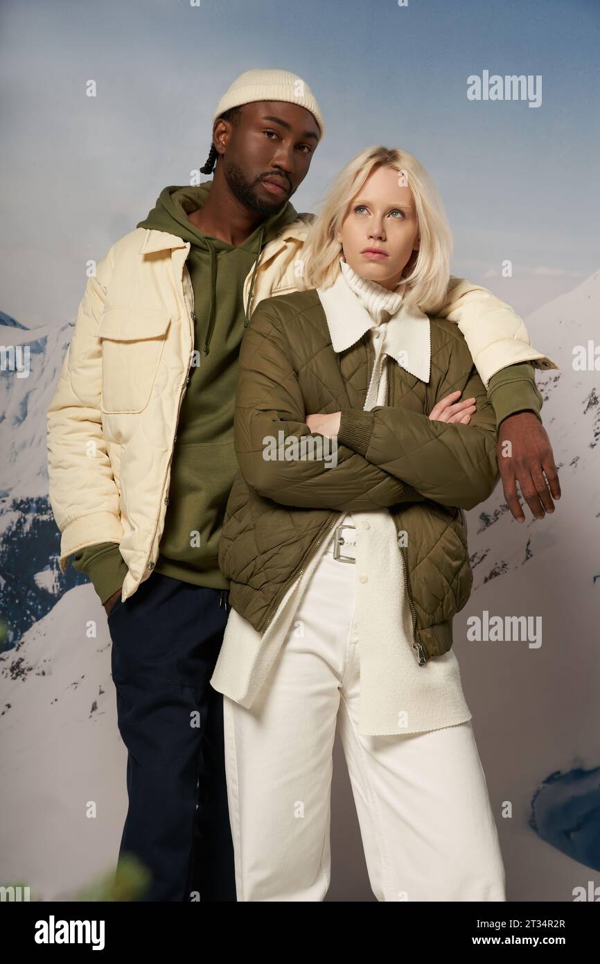 diverse fashionable couple in warm attire posing on snowy backdrop with mountain, winter concept Stock Photo